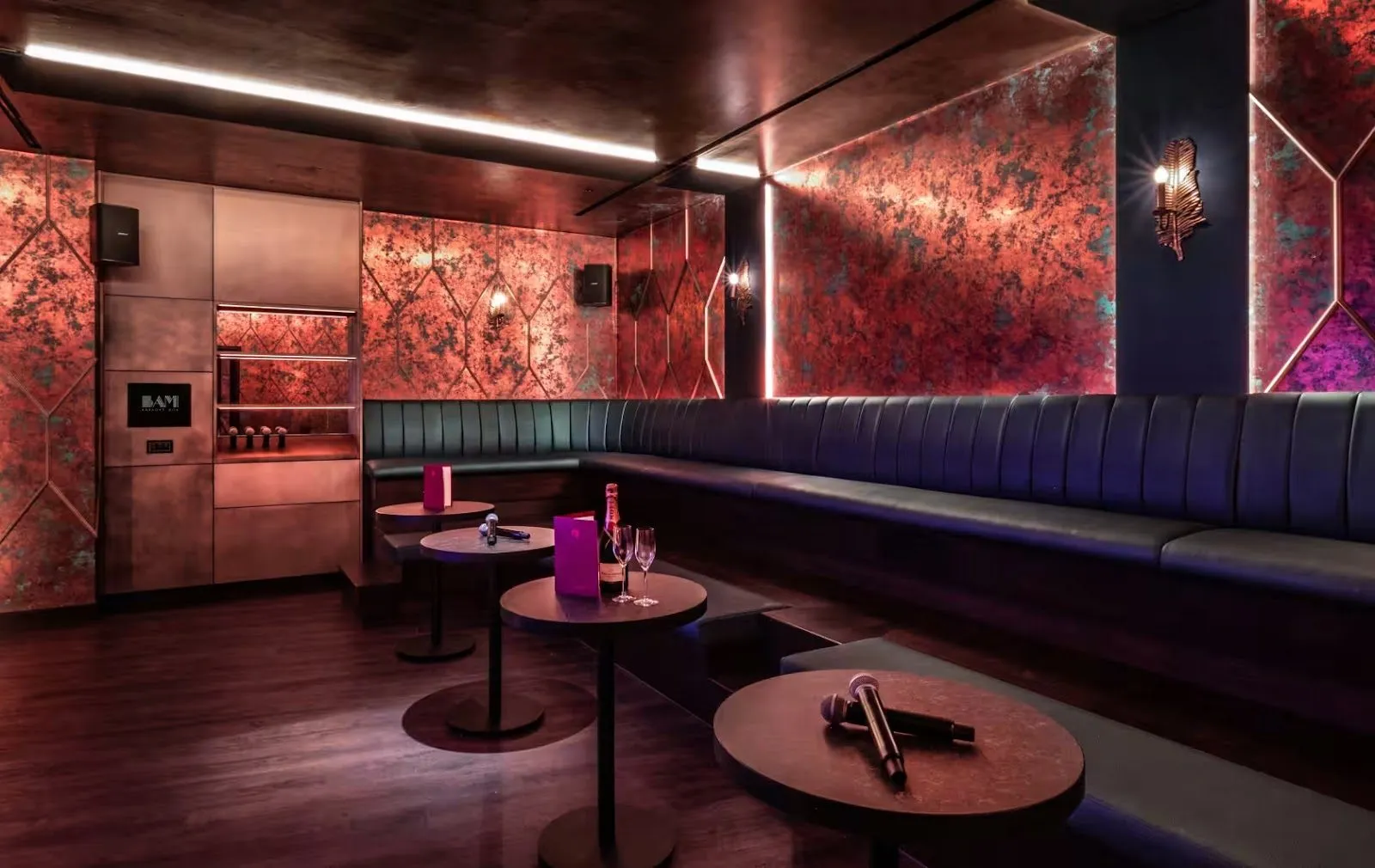A karaoke room at BAM, with a long navy banquette, small round tables with microphones, patterned copper walls and the BAM logo on a screen