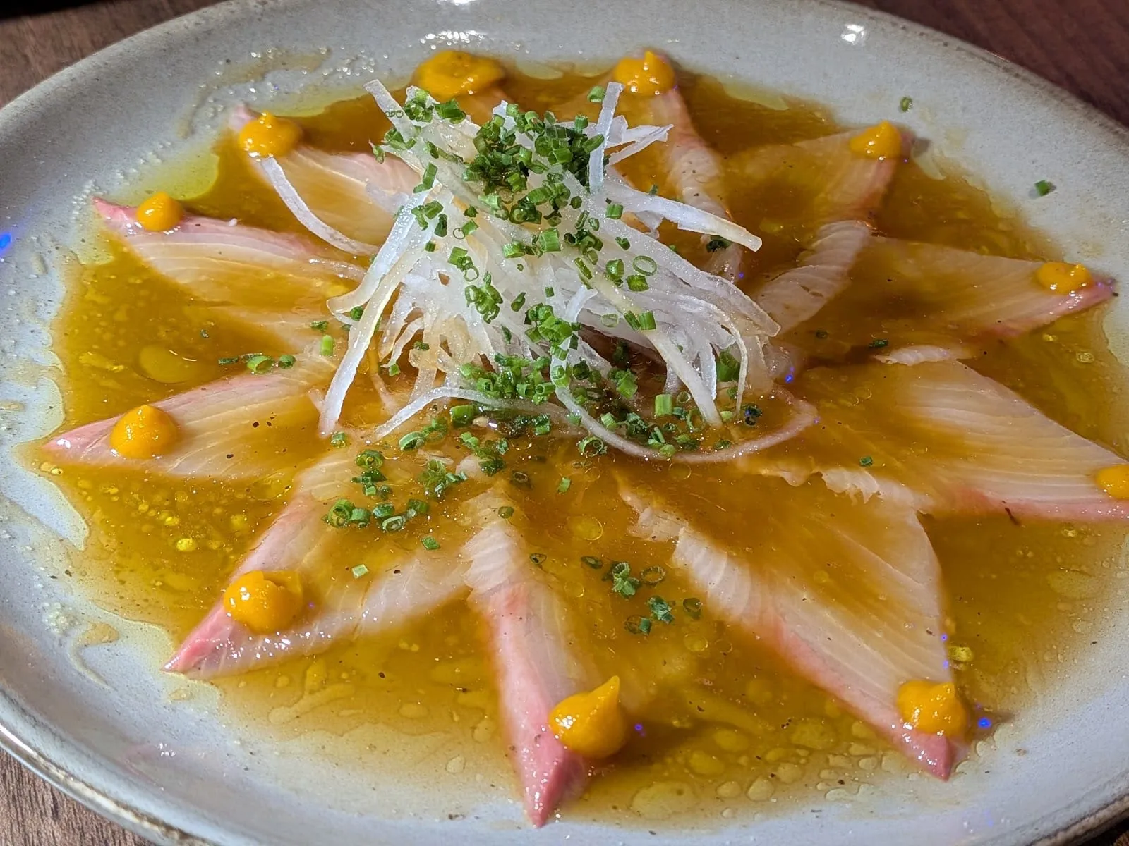 Thin slices of pink-edged yellowtail arranged in a star in a golden marinade, dotted with yellow purée and topped with shredded daikon and chopped chives