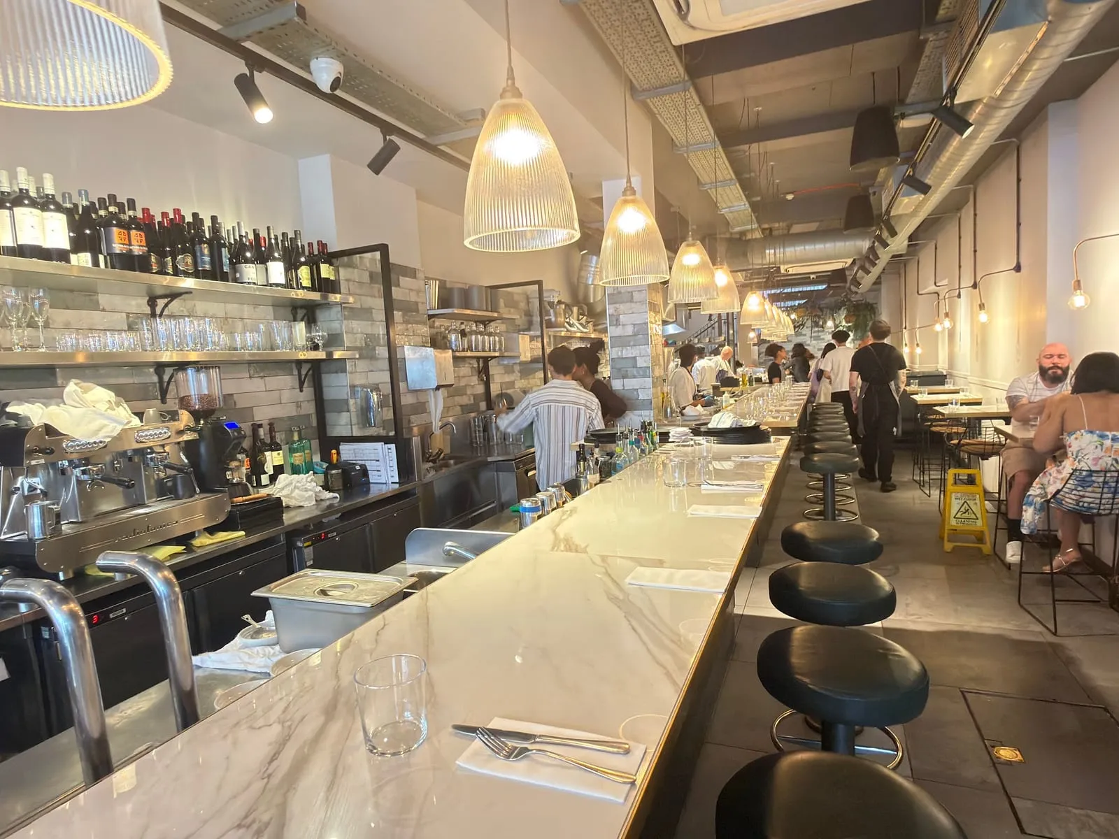 The long marble counter at Bancone, black leather stools along it and pendant lights overhead, with bottles on shelves behind the bar