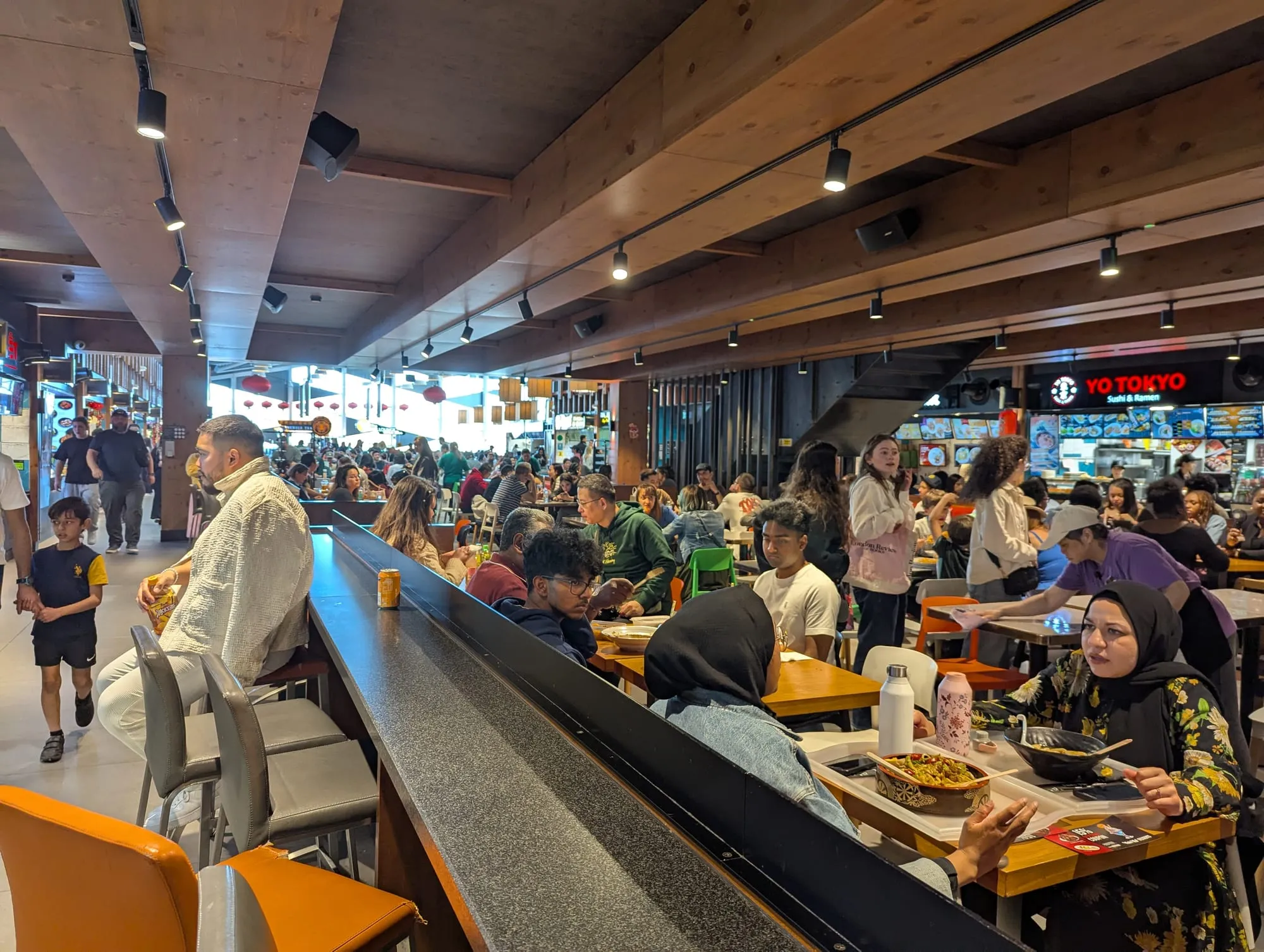 The interior of Bang Bang Oriental, a timber-ceilinged food hall with long communal tables full of people eating, kiosks including a Yo Tokyo sushi and ramen counter along the far wall