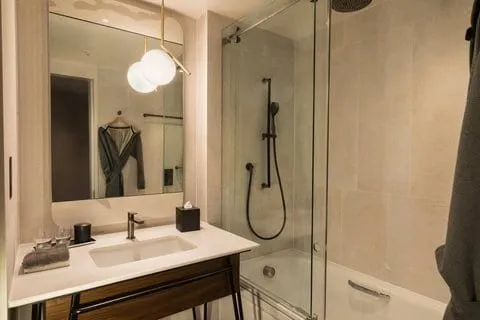 A bathroom at Bankside Hotel with a bath under a glass-screened shower, a pale basin on a wood-and-black-metal stand and globe lights over the mirror