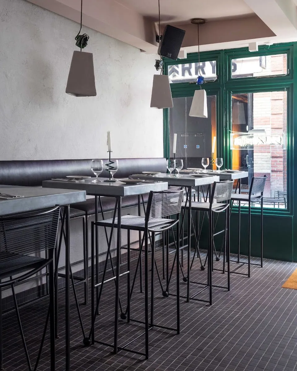 Tall grey tables with black wire bar stools lined up along a banquette, set with wine glasses and candles, beside green-framed windows onto the street