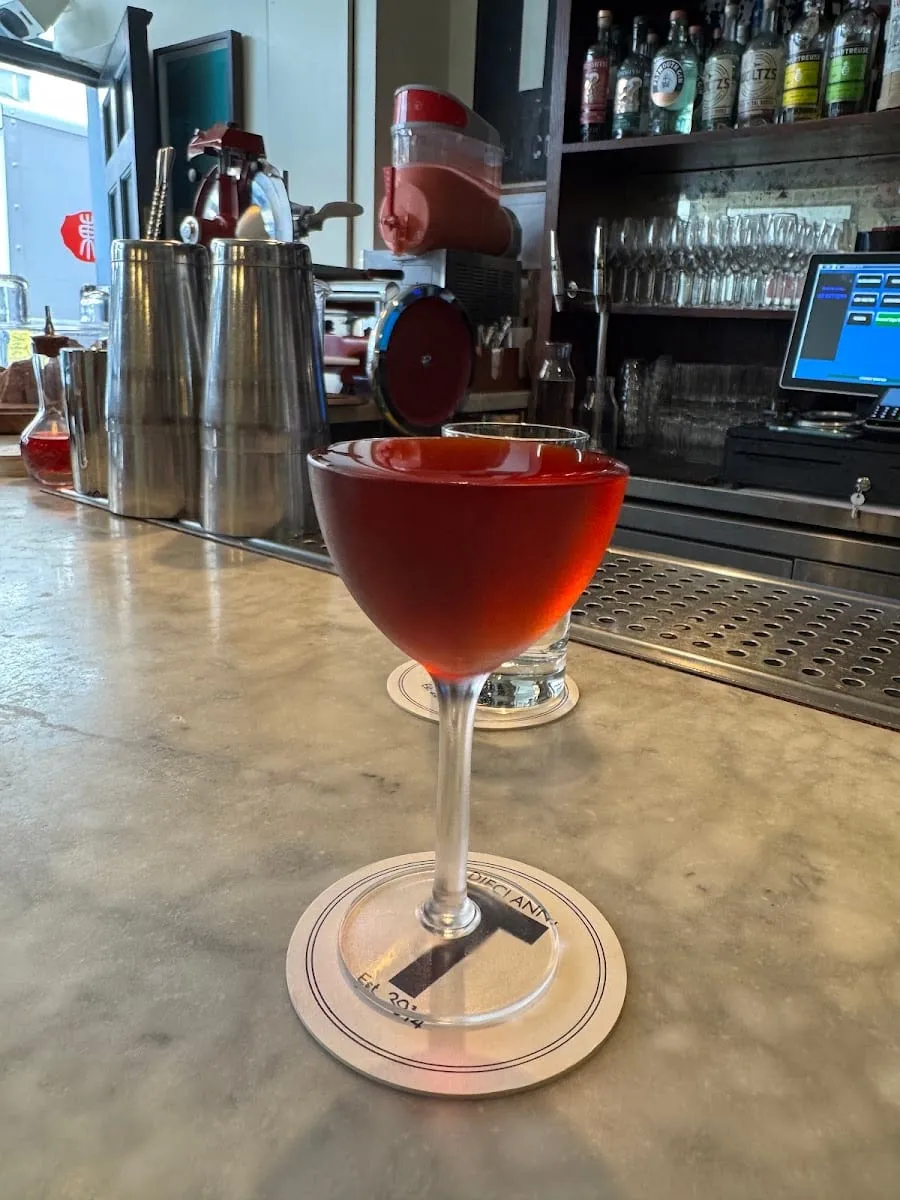A red cocktail served without ice in a small stemmed glass, on a coaster printed with a large T on a marble counter, with steel shakers and shelves of bottles behind