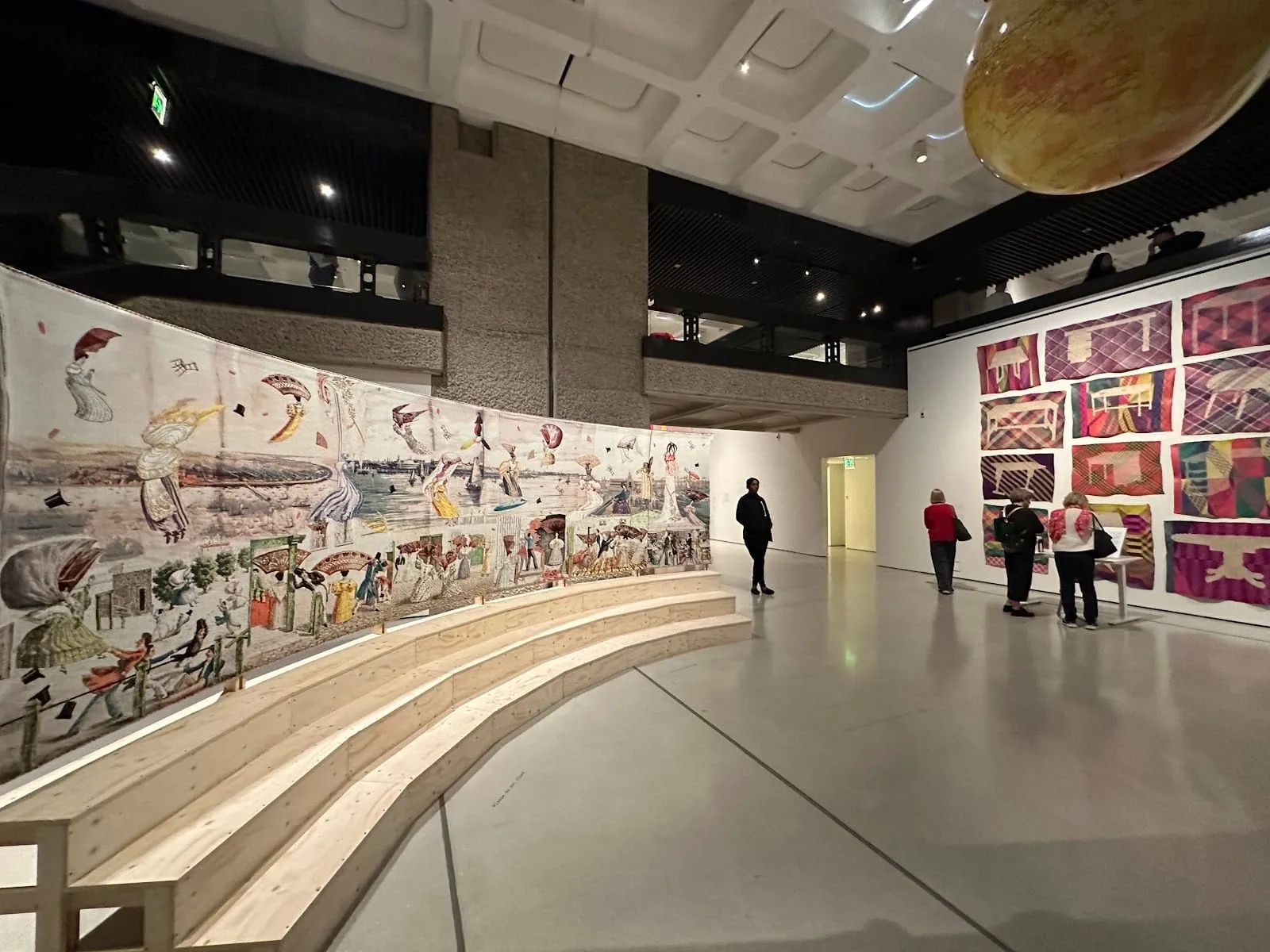 An exhibition in the Barbican Art Gallery: a long illustrated textile hung along curved timber steps, framed textiles on a white wall opposite, and a concrete balcony running round the upper level