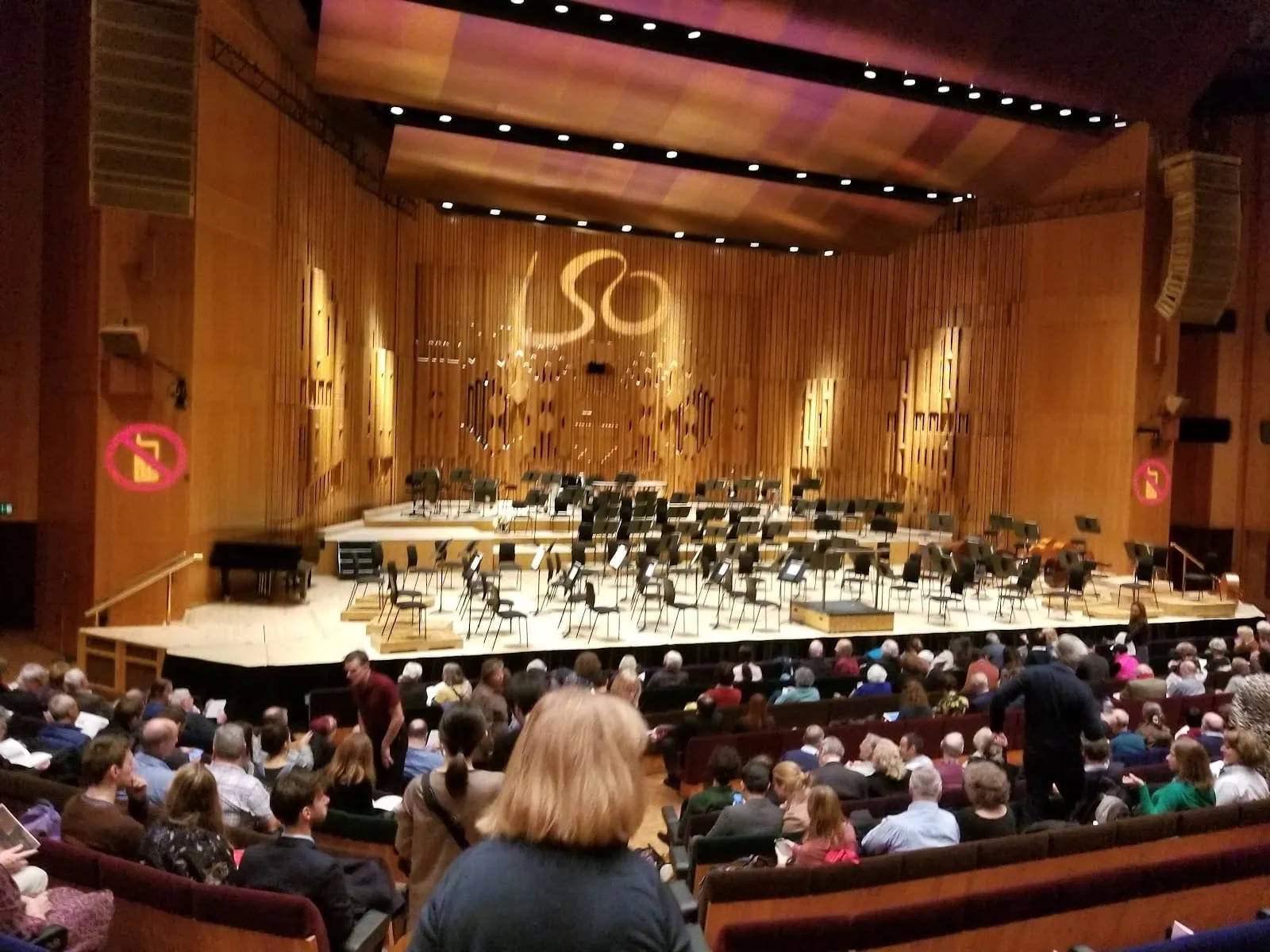 The Barbican Hall from the stalls before a concert: an audience taking its seats, empty chairs and music stands set out for an orchestra, and the LSO logo projected on the wooden wall behind the stage