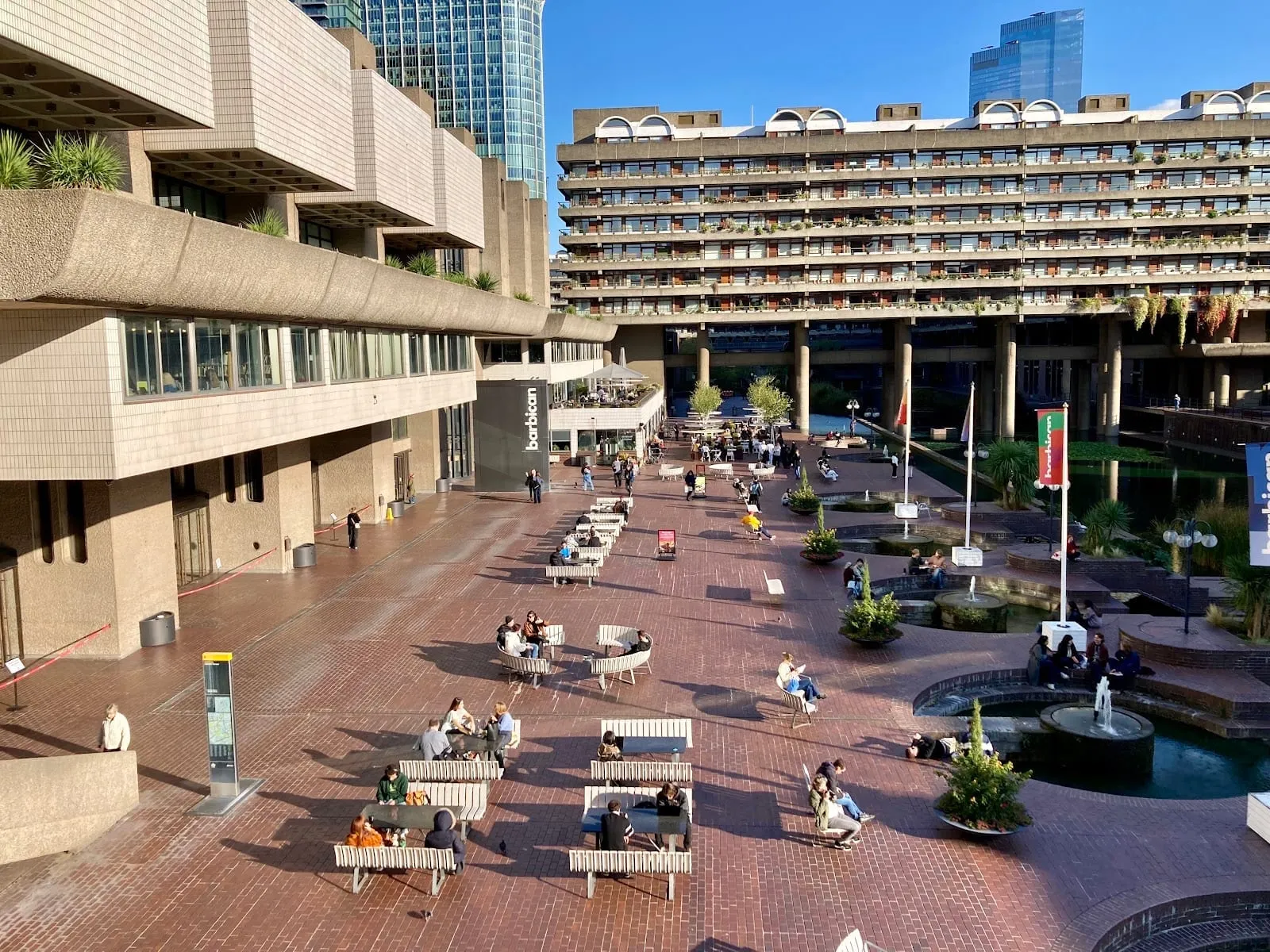 The Barbican Centre's Lakeside Terrace on a sunny day, with people at benches and tables beside the fountains and the long balconied block of Barbican flats behind