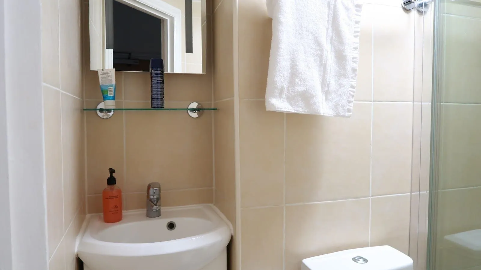A small beige-tiled en-suite at Barry House with a compact basin, a glass shelf under a mirror and a glass shower door