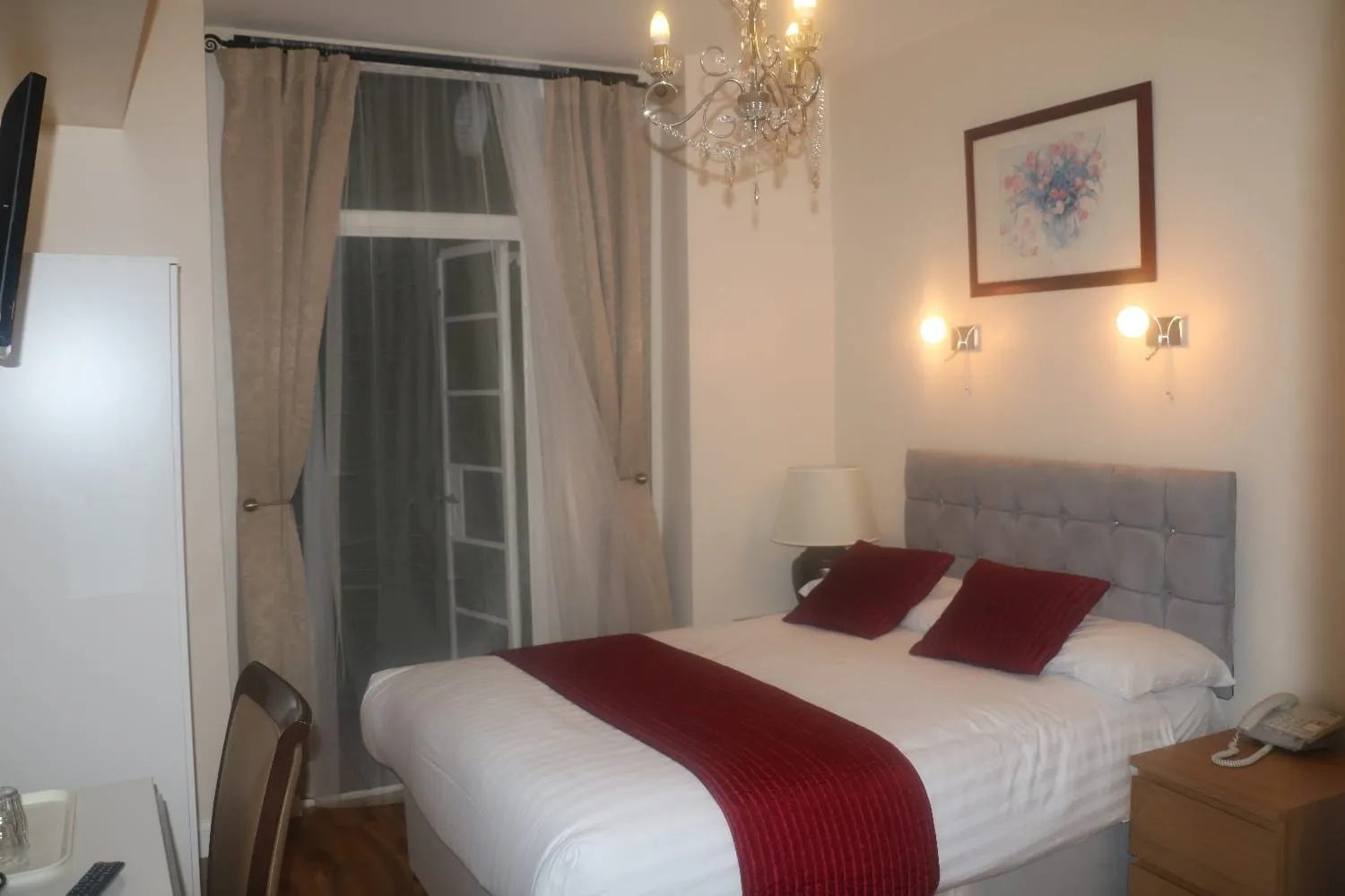 A double room at Barry House with a grey buttoned headboard, a red bed runner, a small crystal chandelier and full-height French windows behind voile curtains