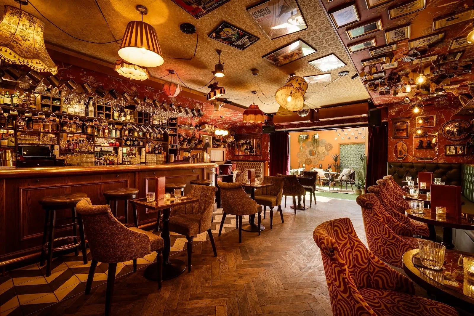 The bar room at Barts, with fringed lampshades, framed pictures covering the ceiling, patterned armchairs and a doorway to a garden at the back