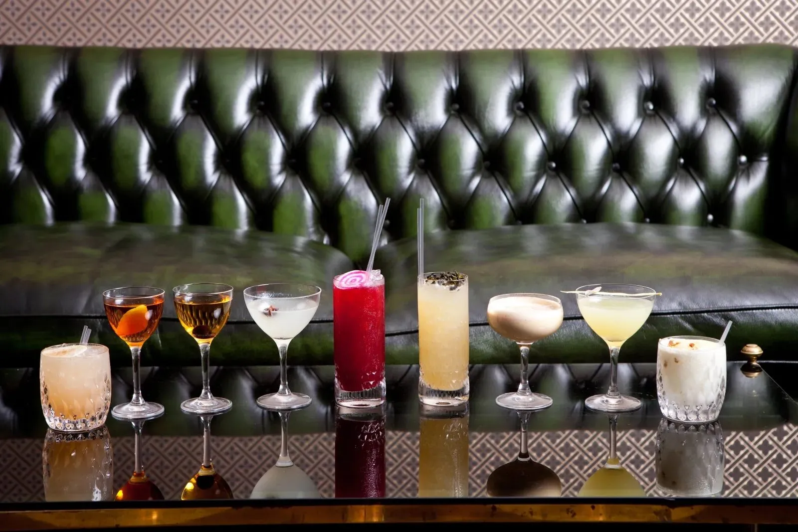 A row of cocktails on a glass table in front of a green buttoned-leather sofa at Basement Sate