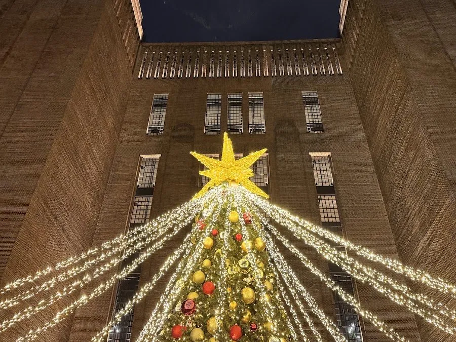 A Christmas tree hung with gold and red baubles and topped with a gold star, with strands of lights fanning out from it against the brick wall and tall windows of Battersea Power Station at night