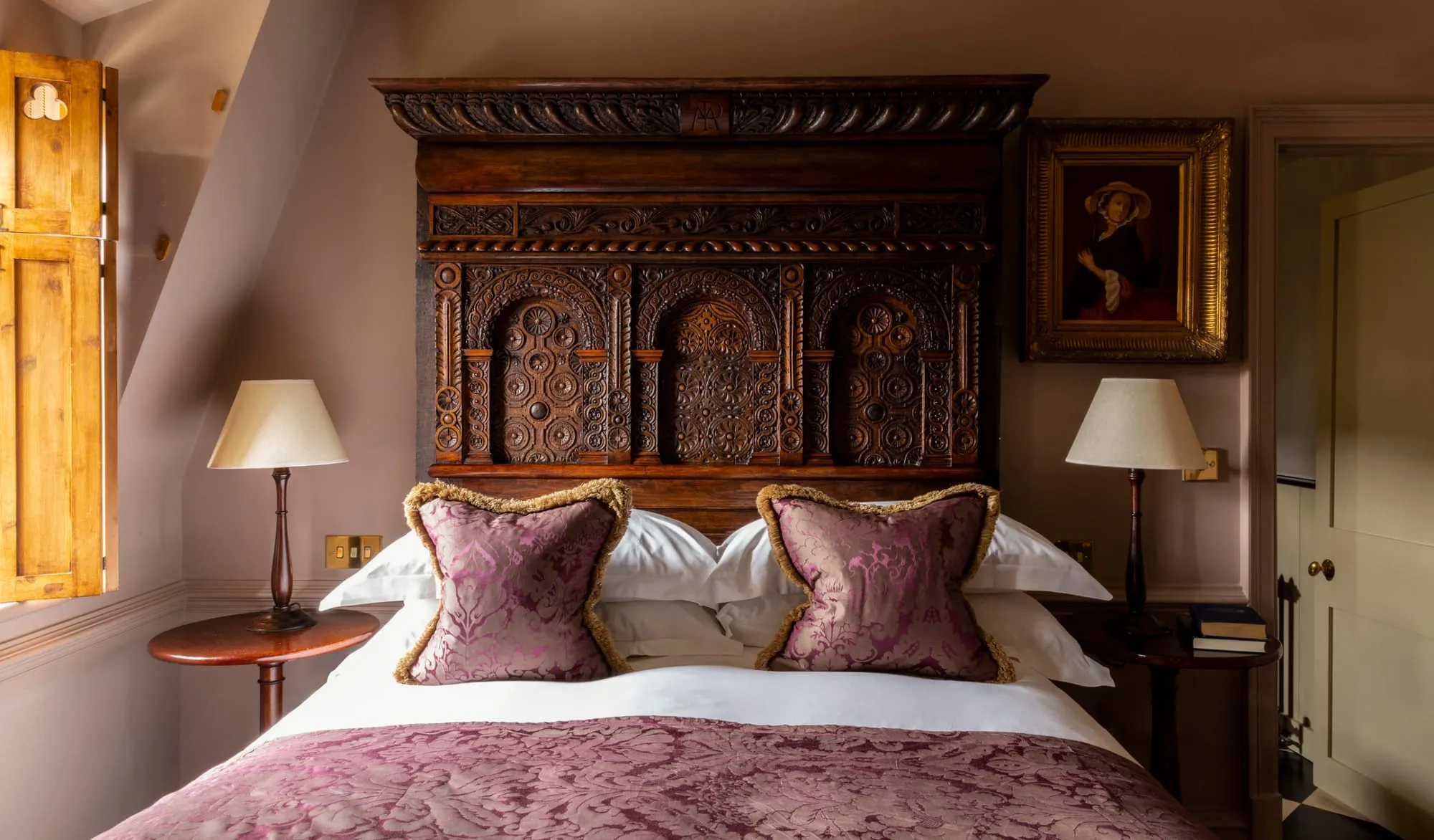 A Batty Langley's bedroom with an ornately carved dark wood four-poster headboard, an antique oil portrait on the wall, and purple damask cushions