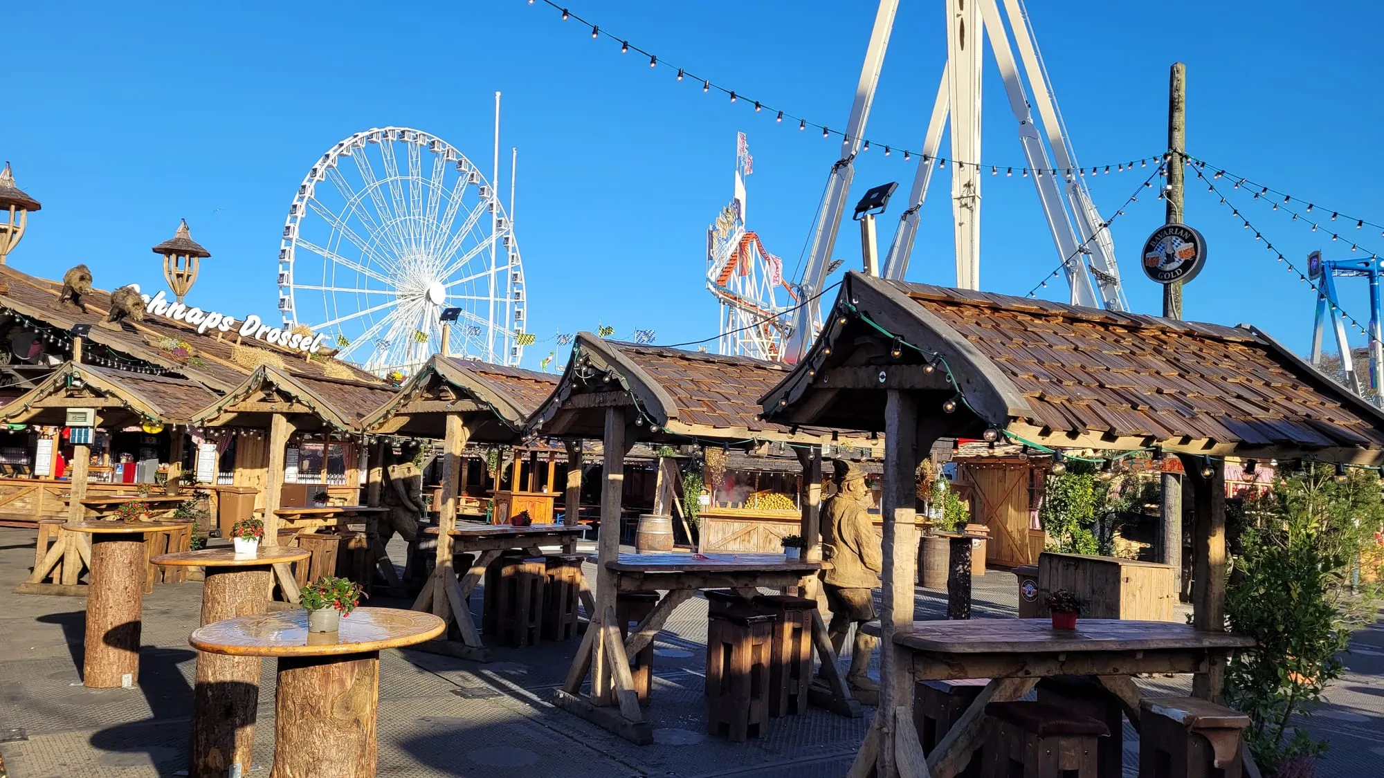 Wooden Bavarian-style stalls at Winter Wonderland in bright winter sunshine, with the observation wheel and a rollercoaster behind