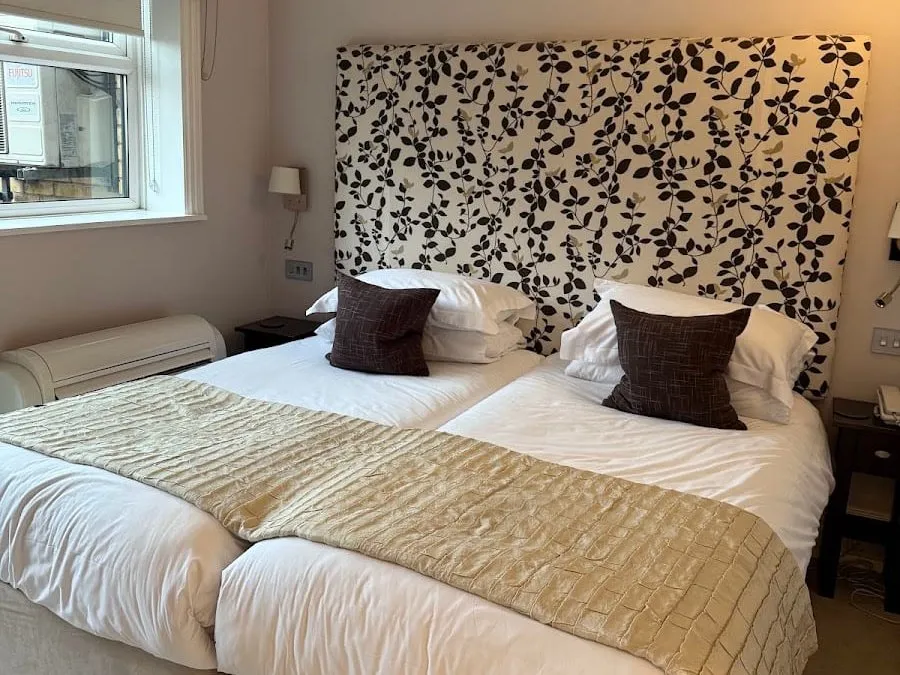 A bedroom at Beaufort House with two single beds pushed together under a tall black-and-white leaf-print headboard, a gold throw, and a window looking onto air-conditioning units