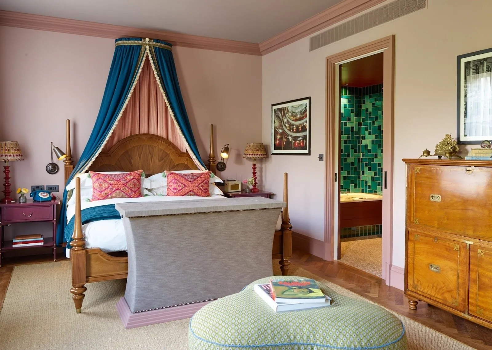 A bedroom at Beaverbrook Town House: a four-poster bed under a blue and pink canopy, dusty-pink walls, a campaign chest, a photograph of a theatre auditorium, and a doorway to a bathroom tiled in green