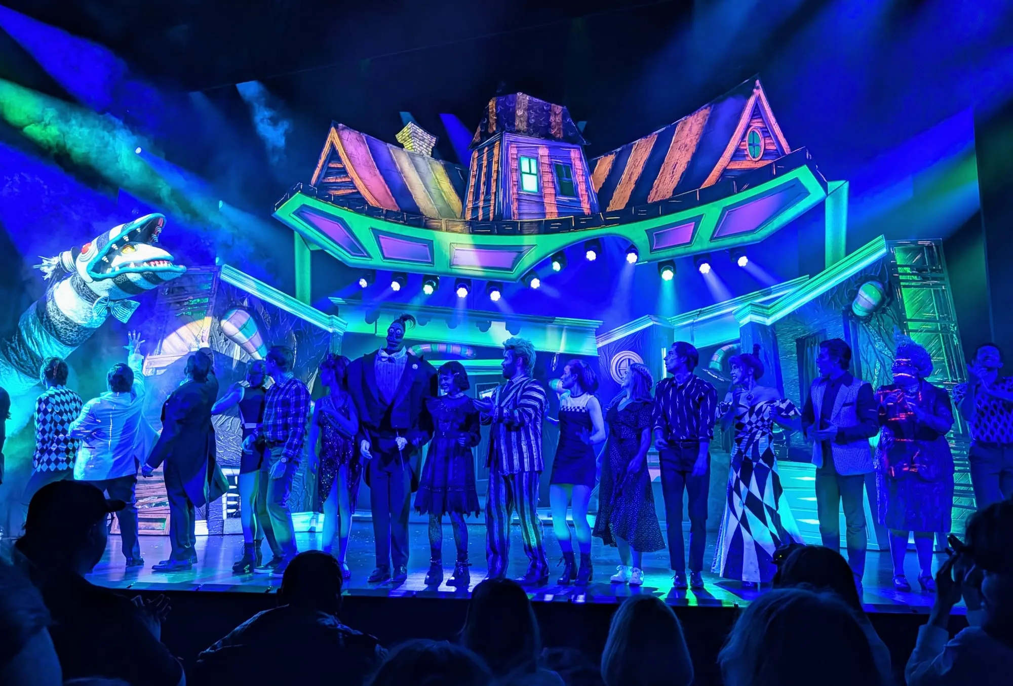 The cast of Beetlejuice taking their curtain call at the Prince Edward Theatre, lit in green and blue, with the Maitland house tilting above them and the sandworm reared up at the side of the stage