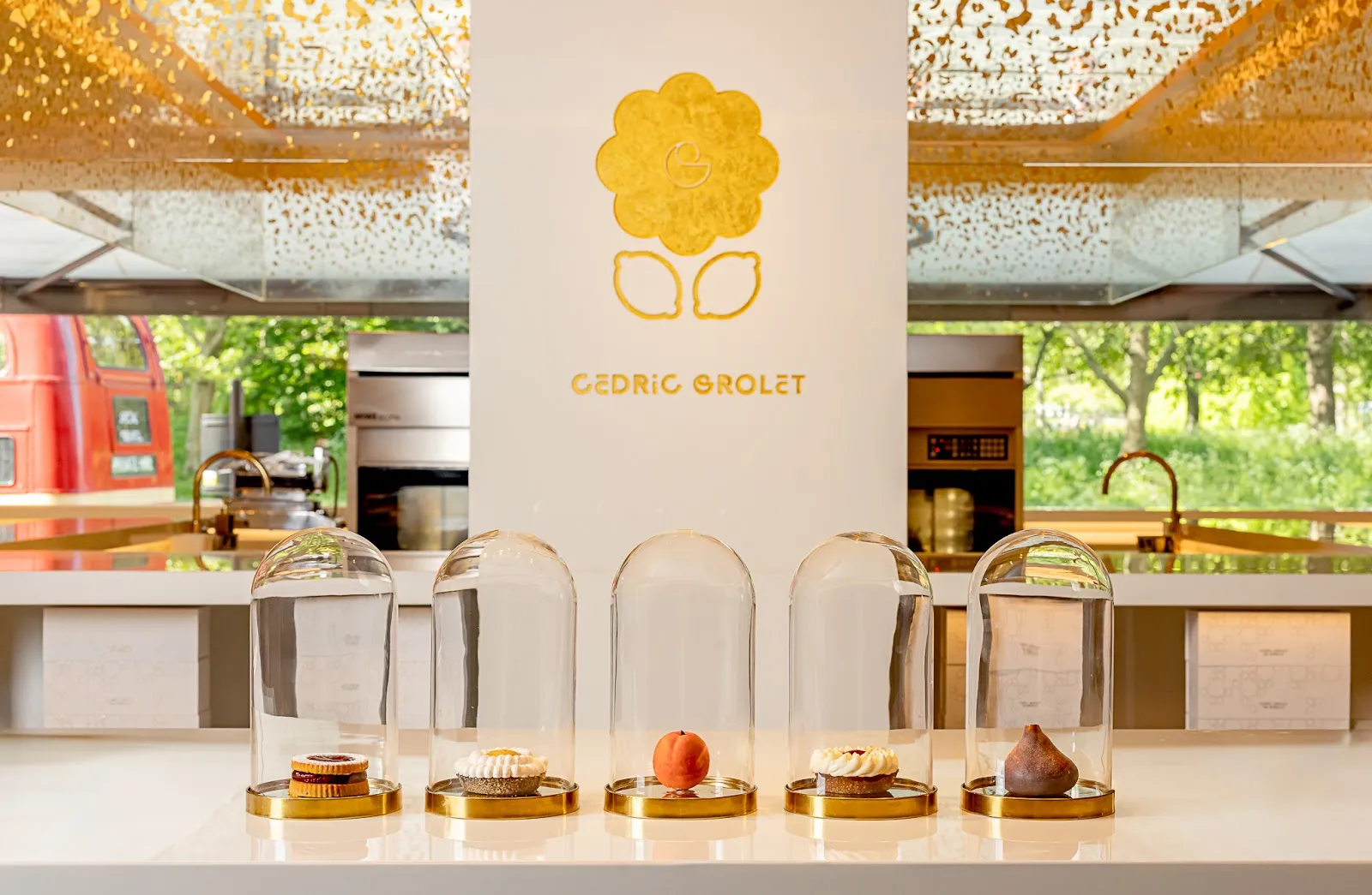 Five pastries under glass cloches on the Cedric Grolet counter at The Berkeley, including a trompe-l'oeil peach and fig, with Hyde Park through the window behind