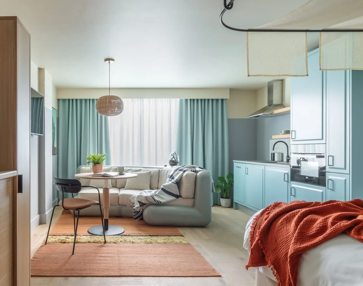 A studio at Bermonds Locke, with a sofa and dining table beside the window and a pale blue kitchenette along the back wall, the bed in the foreground with a rust-coloured throw