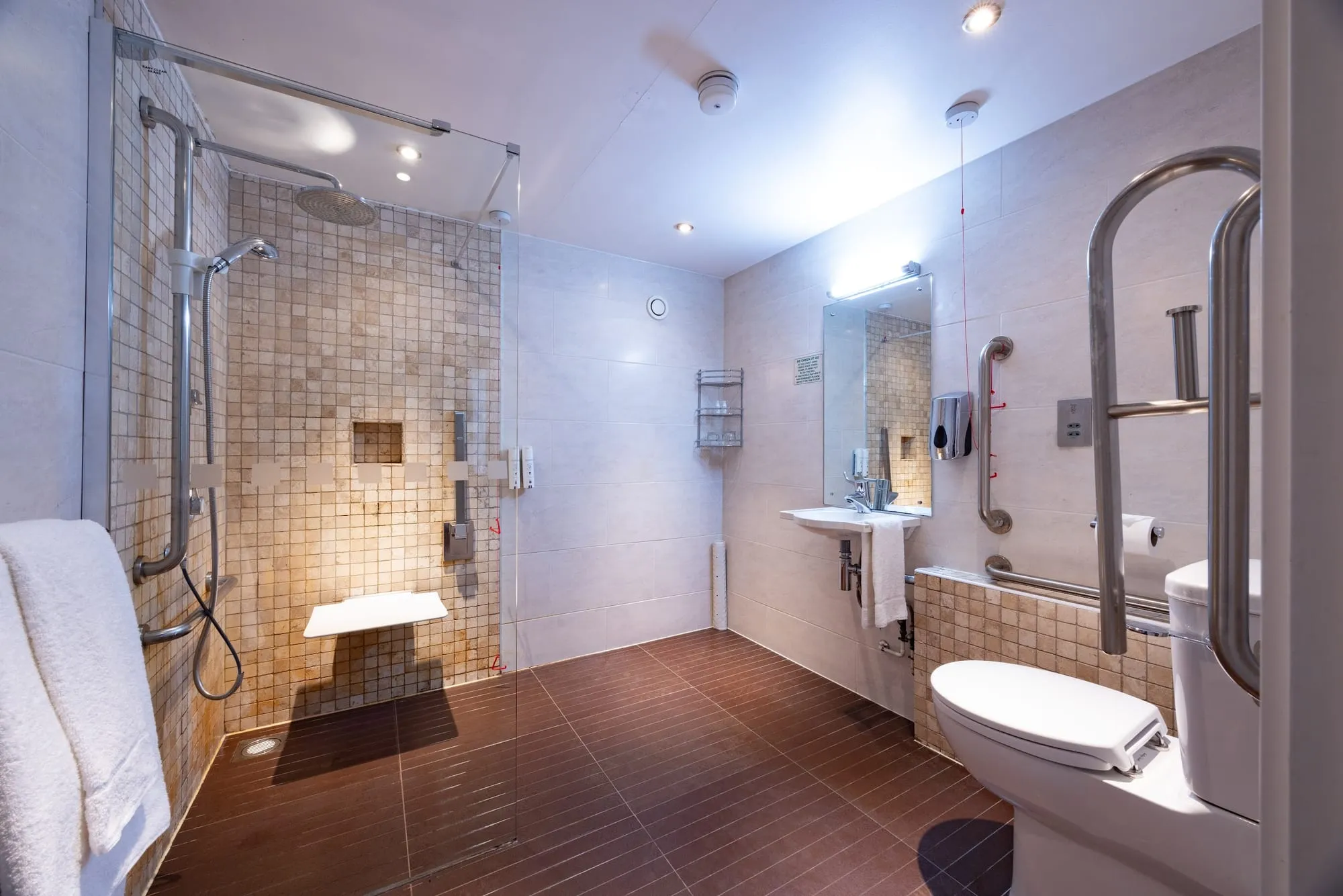 The bathroom in a room at Bermondsey Square Hotel, finished in travertine-look tile with a walk-in shower and grab rails
