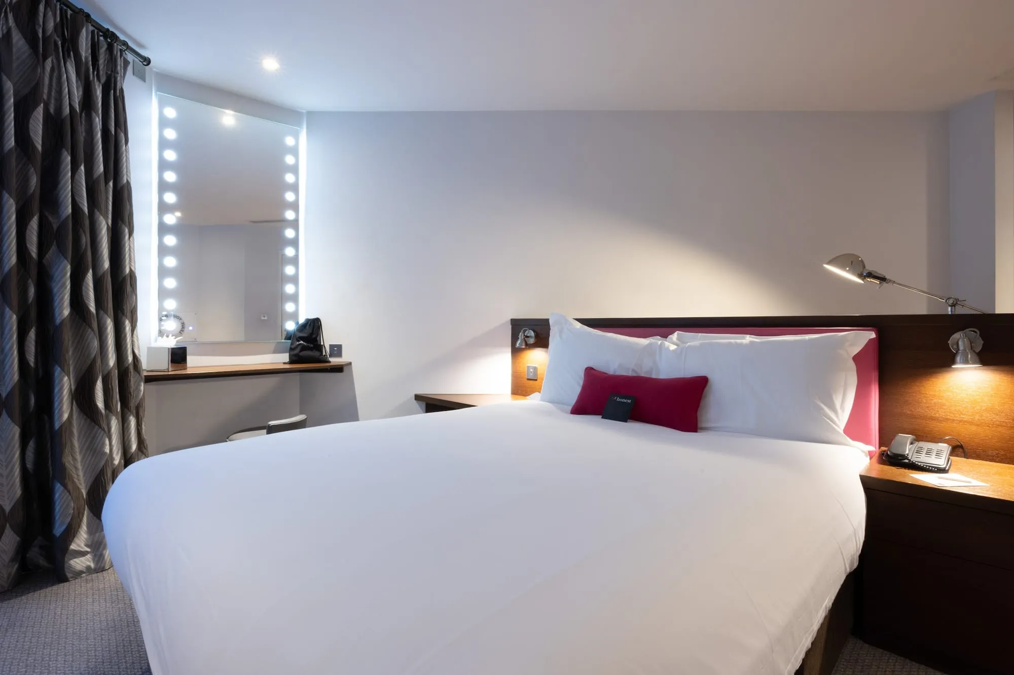 A Bermondsey Square Hotel room with patterned curtains, an illuminated vanity mirror and a pink accent pillow on the bed