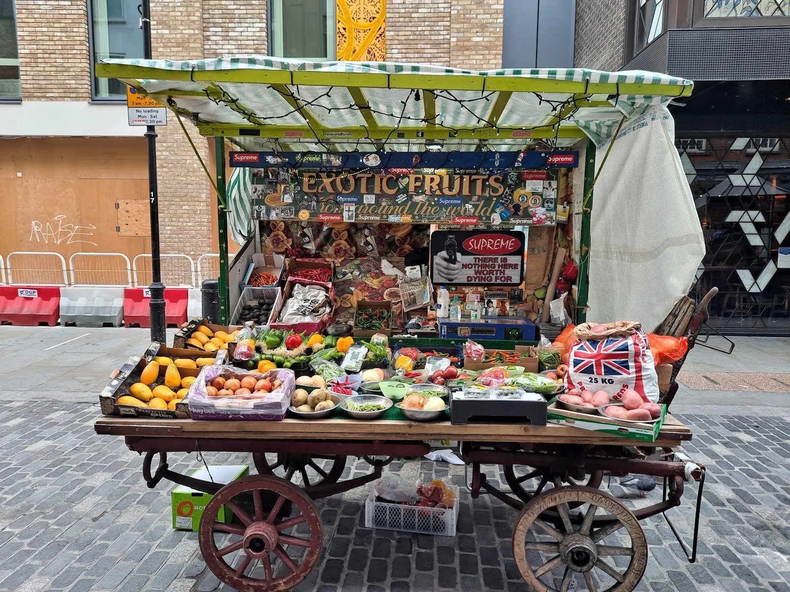 A fruit and vegetable barrow on wooden cartwheels on Berwick Street, under a striped awning and an Exotic Fruits sign, piled with papayas, peppers, chillies and a sack of potatoes