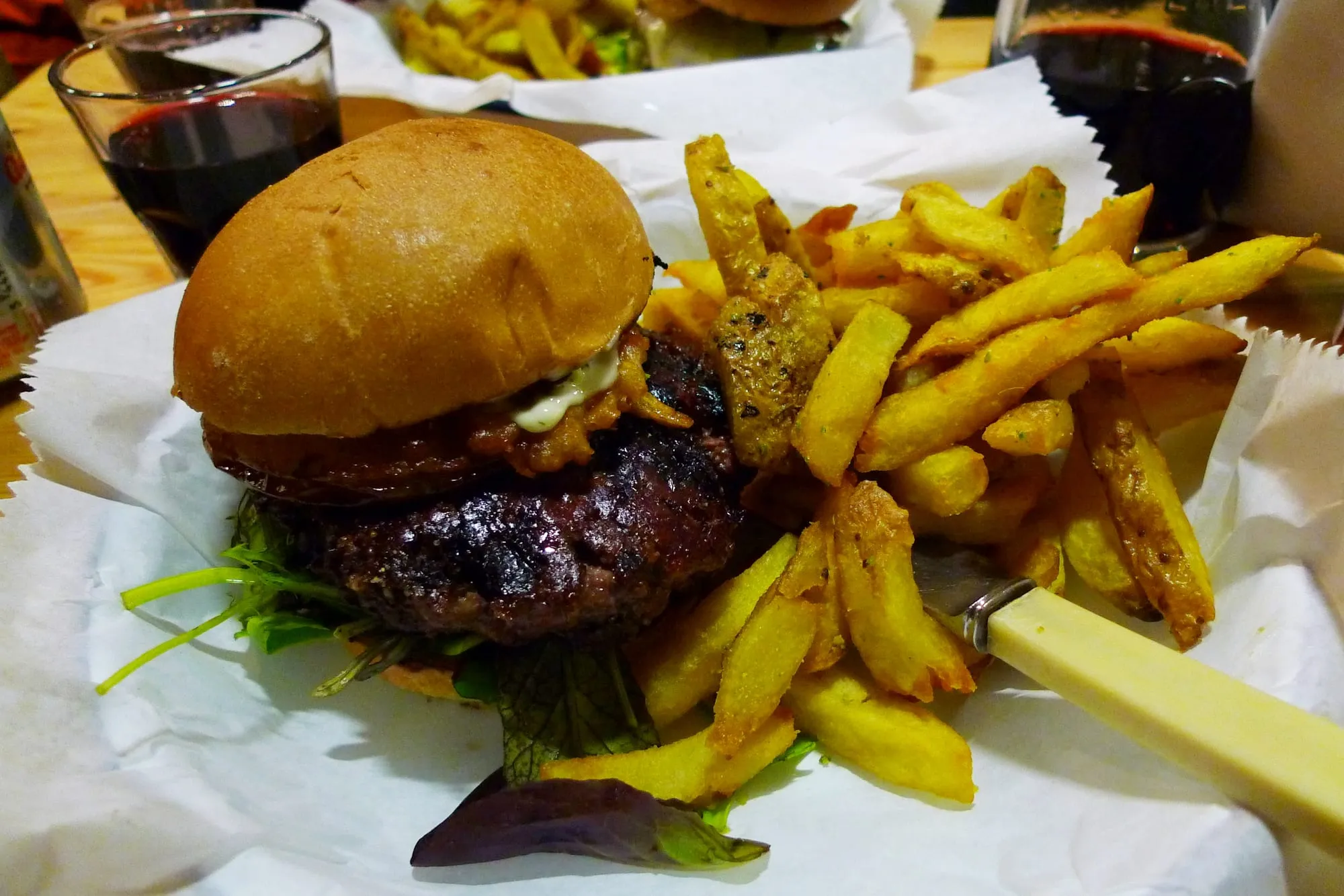 A thick charred beef patty in a soft bun beside a pile of rosemary-salted chips and a glass of red wine
