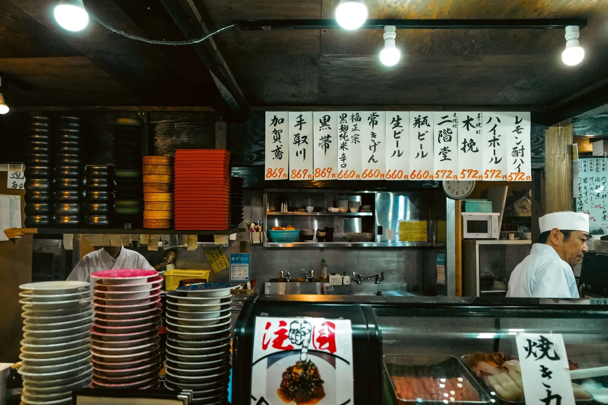 The counter of a Japanese restaurant with stacked plates and handwritten menu boards
