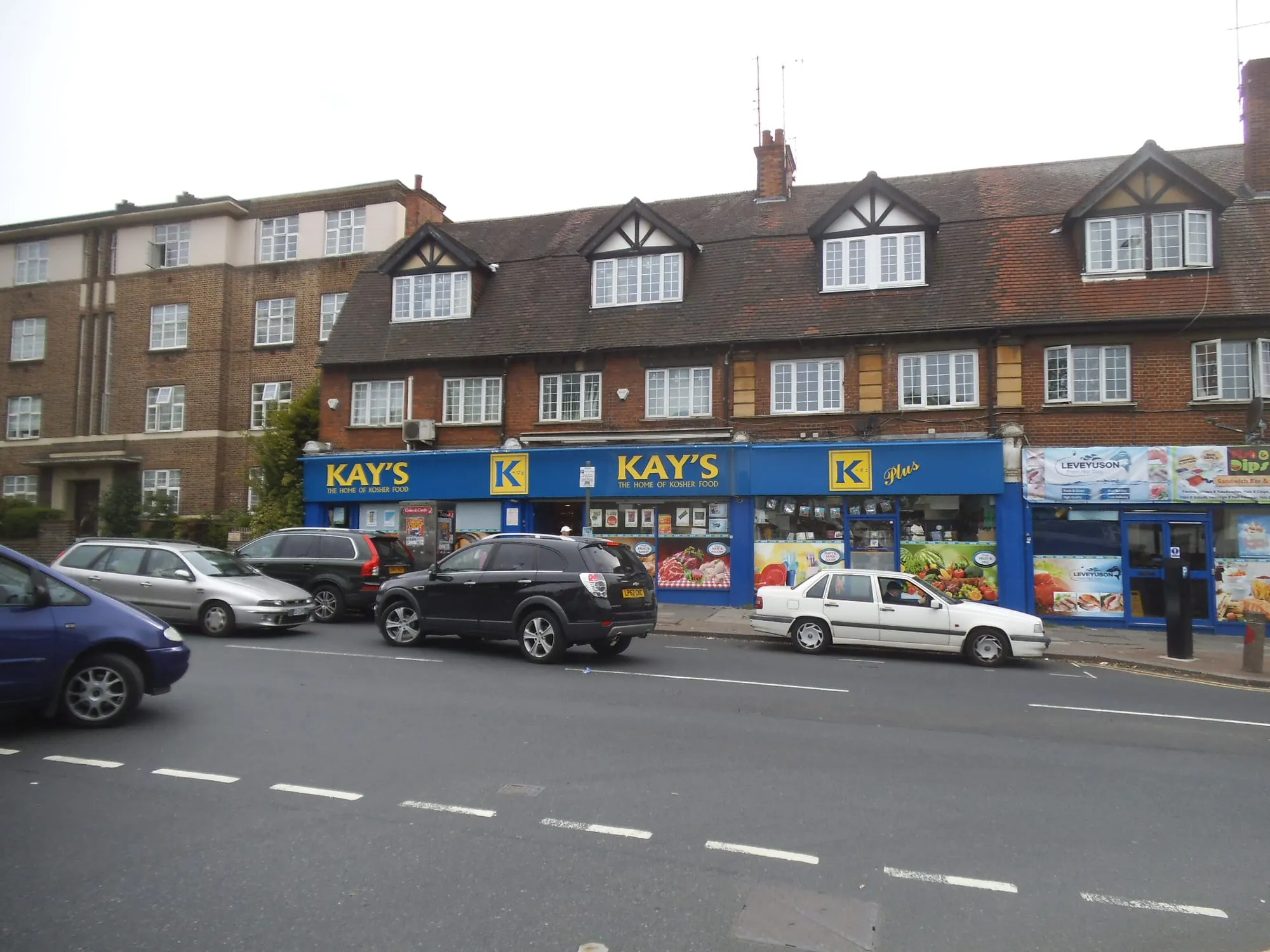 A parade of kosher shops on Golders Green Road - Kay's, signed 'the home of kosher food', with Kay's Plus and the Leveyuson fishmonger alongside - below Tudor-revival flats, with traffic passing on the road in front