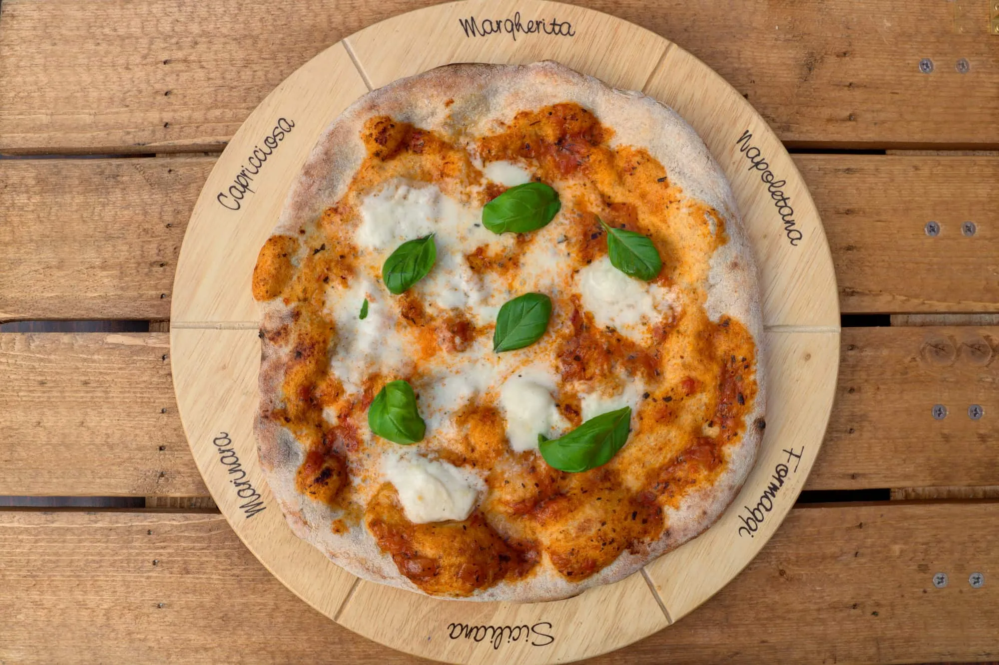 A margherita pizza on a wooden board marked with the names of classic pizza styles — Margherita, Napoletana, Formaggi, Siciliana, Marinara and Capricciosa