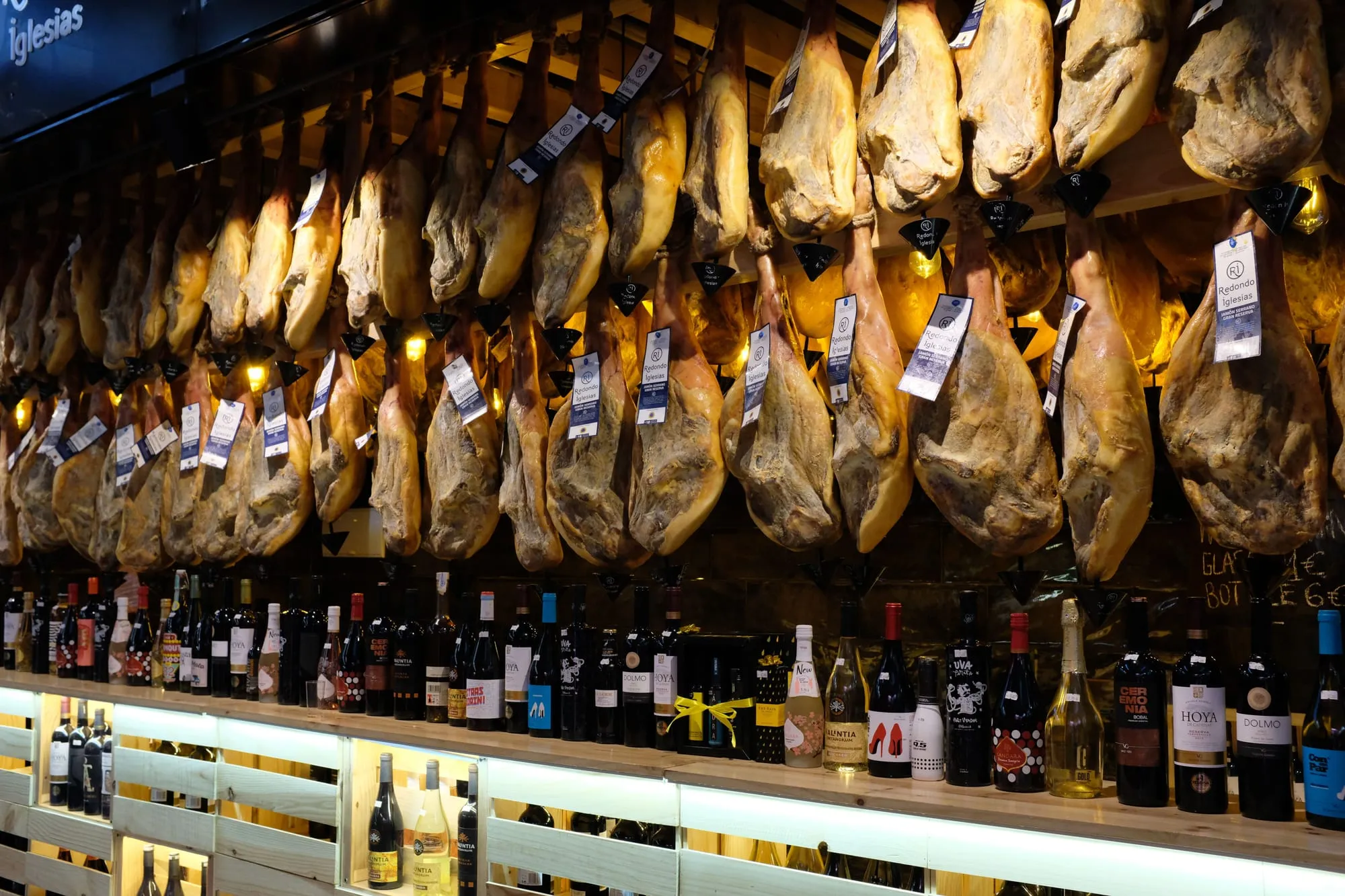 Whole jamón legs hanging above a counter lined with wine bottles