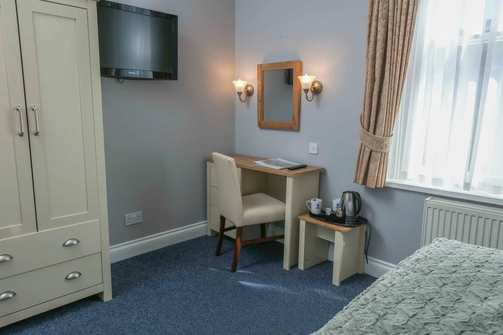 A room at the Best Western Swiss Cottage with lilac-grey walls, a cream Shaker-style wardrobe, a small desk under a wood-framed mirror between two brass wall lights, a kettle tray and a tall window with gold curtains