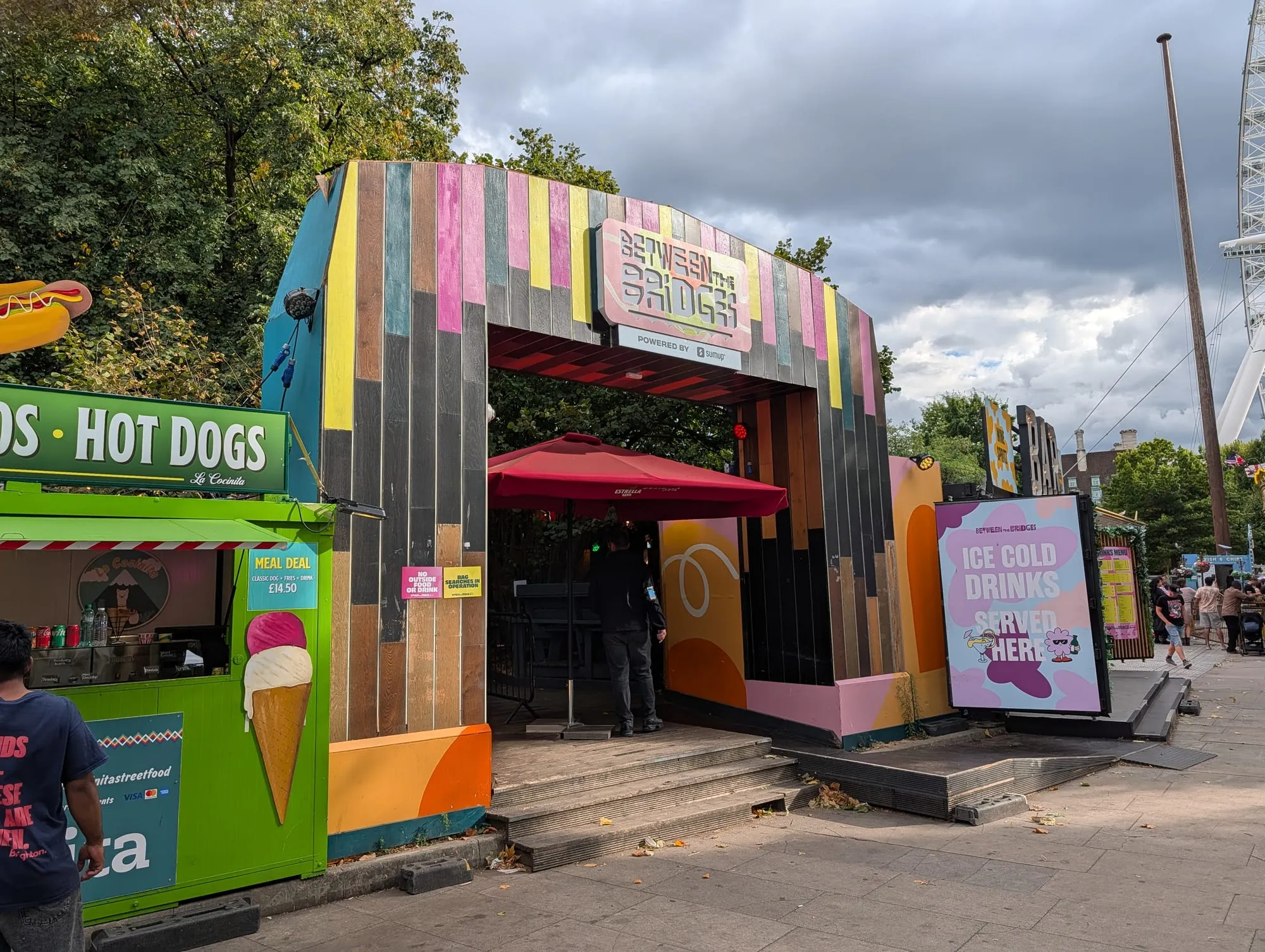 The painted timber entrance arch to Between the Bridges on the South Bank, a hot dog stall to one side and an ice cold drinks board to the other, with the London Eye behind