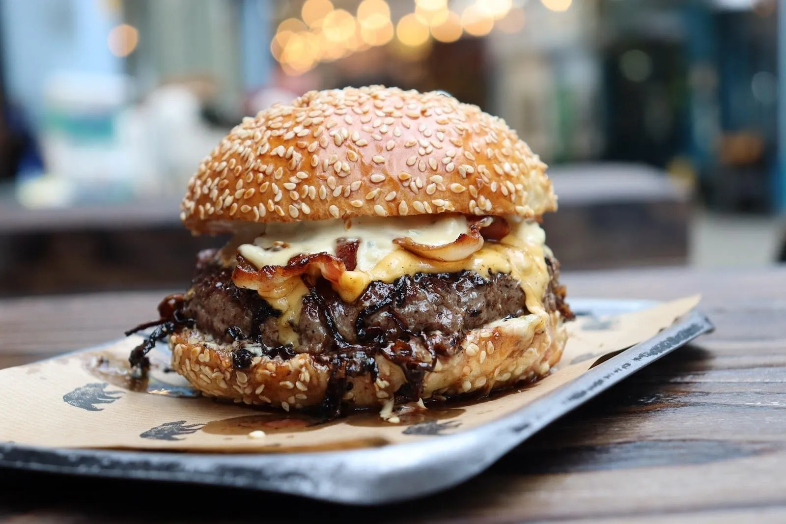 A Black Bear burger on a metal tray lined with bear-print paper: a thick beef patty with dark caramelised onions, melted cheese, bacon and a pale mayo in a sesame-crusted bun