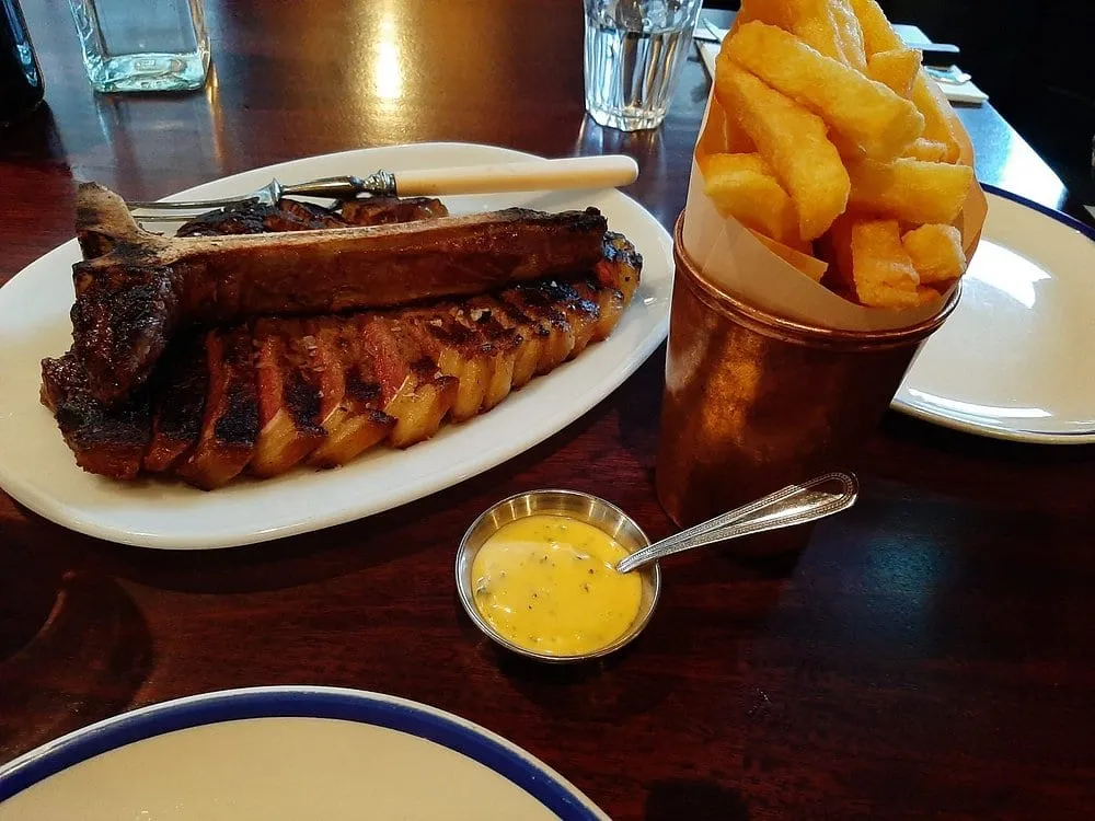 A sliced bone-in T-bone on a white oval plate in a dark room, beside a copper cup of thick-cut chips and a small metal pot of béarnaise