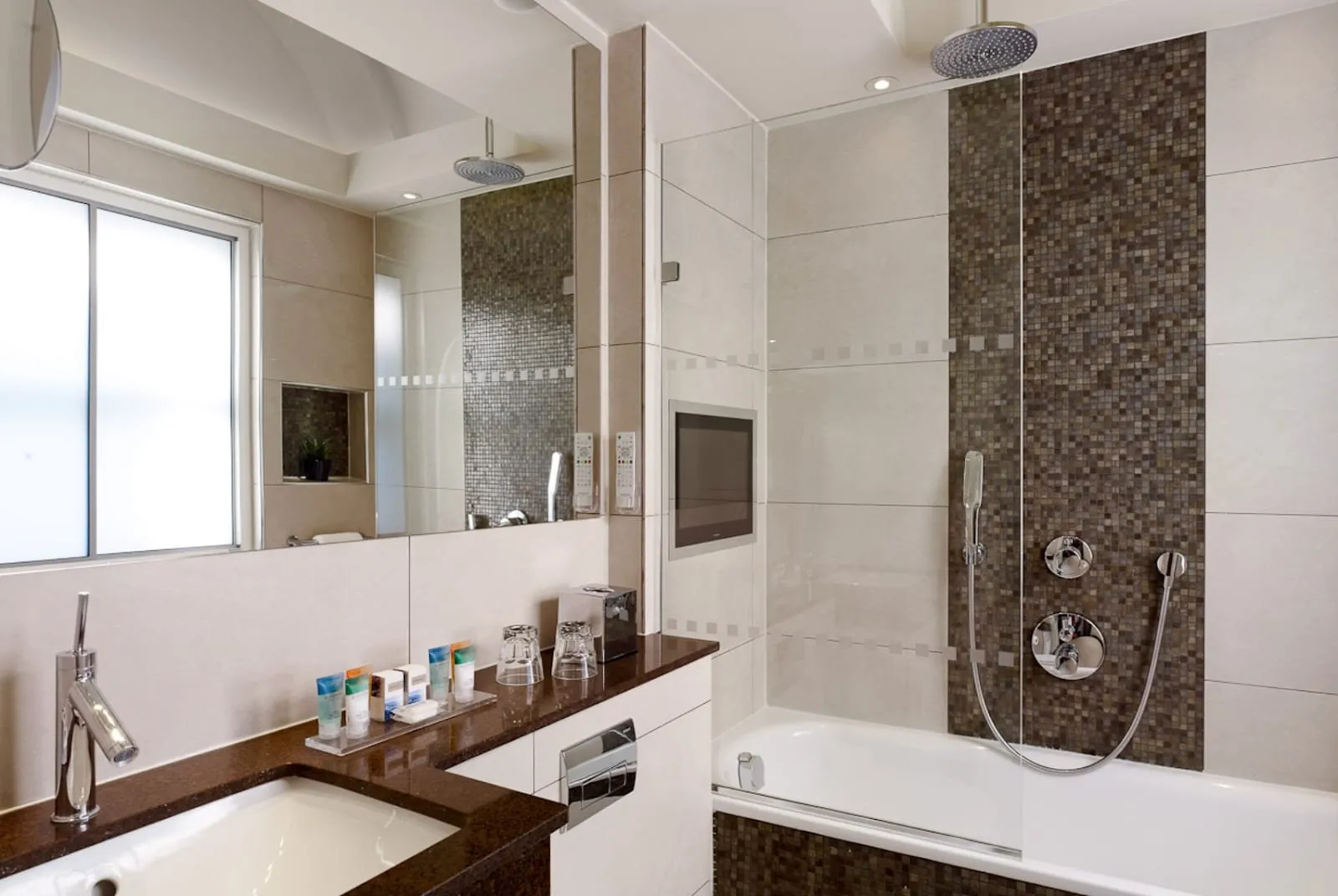 A bathroom at Blakemore Hyde Park with cream tiles, a bronze mosaic stripe, a bath with a rain shower and glass screen, a TV set into the wall and a basin in a dark granite counter
