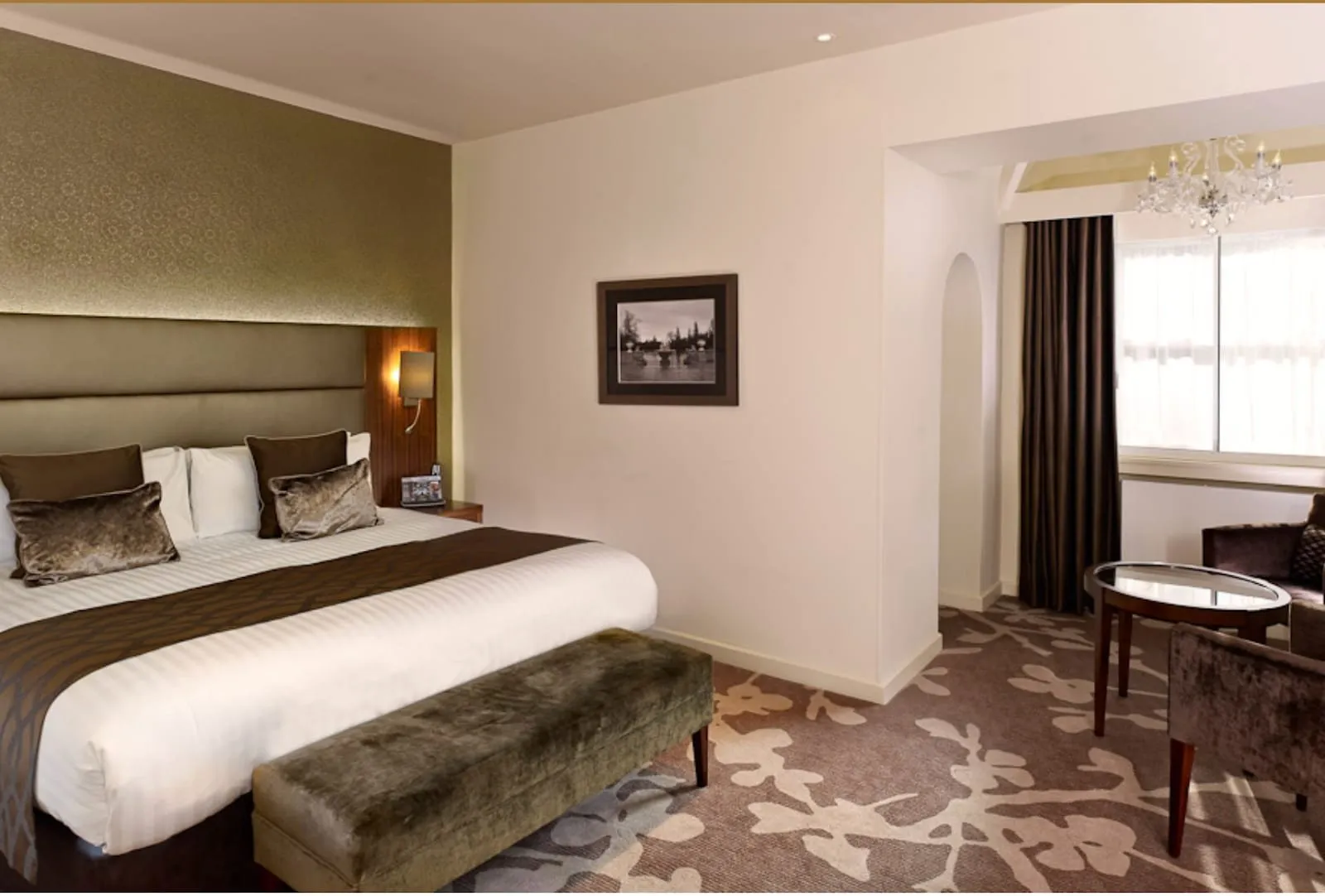 A room at Blakemore Hyde Park with a gold-patterned feature wall, a large bed with velvet cushions and a velvet bench at its foot, and a separate seating area with a round glass table and armchairs under a chandelier by the window