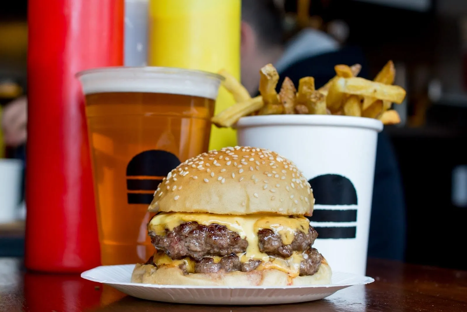 A Bleecker double cheeseburger in a sesame bun on a paper plate, two seared patties under melted American cheese, with a pint of lager and a paper tub of skin-on fries behind it
