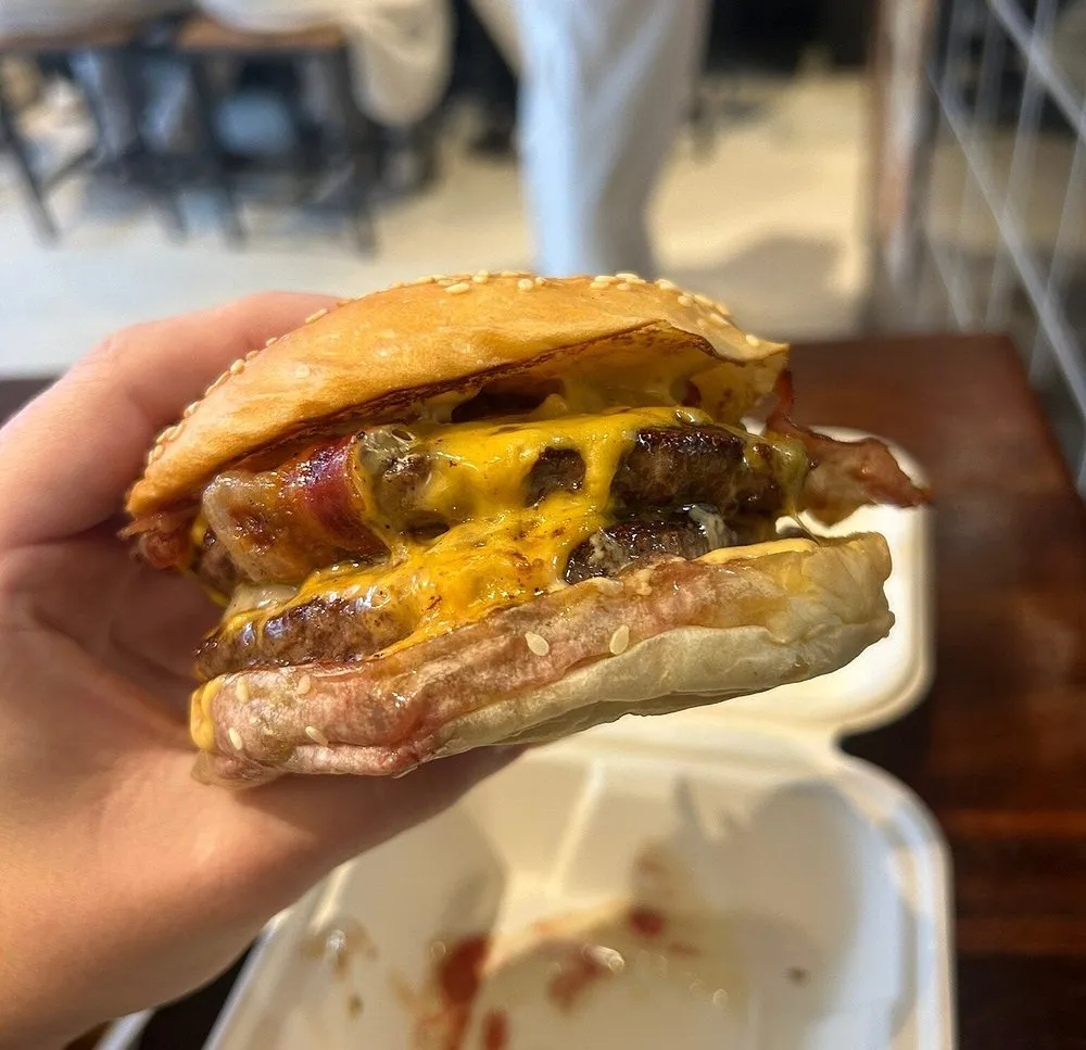 A double cheeseburger with bacon and melted cheese in a sesame bun, held in one hand over an open takeaway box