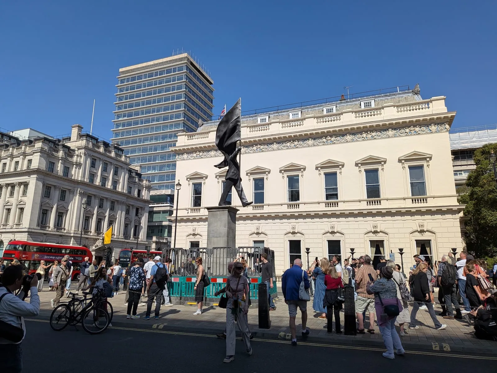 Banksy's Blind Patriotism statue on its plinth on Waterloo Place, a flag blowing across the figure's face, with a crowd photographing it from behind green barriers