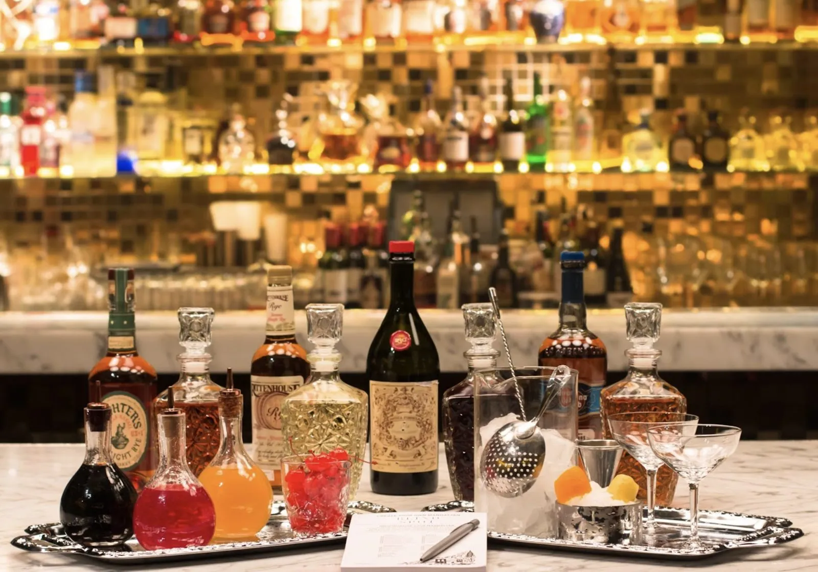 Decanters, bottles, cocktail cherries and a mixing glass on silver trays on a marble bar, in front of shelves of bottles lit gold