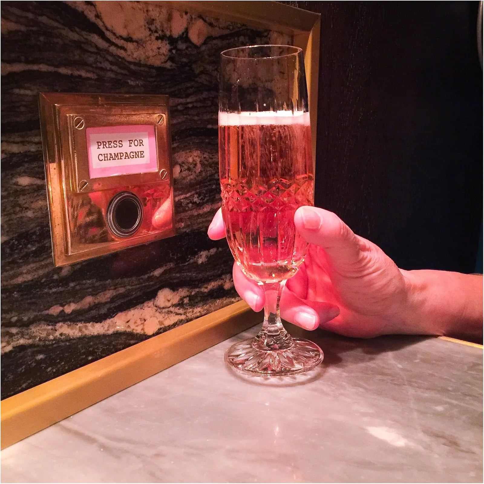 The press-for-champagne button in a brass plate on a marble wall at Bob Bob Ricard, with a hand holding a glass of rosé champagne beside it
