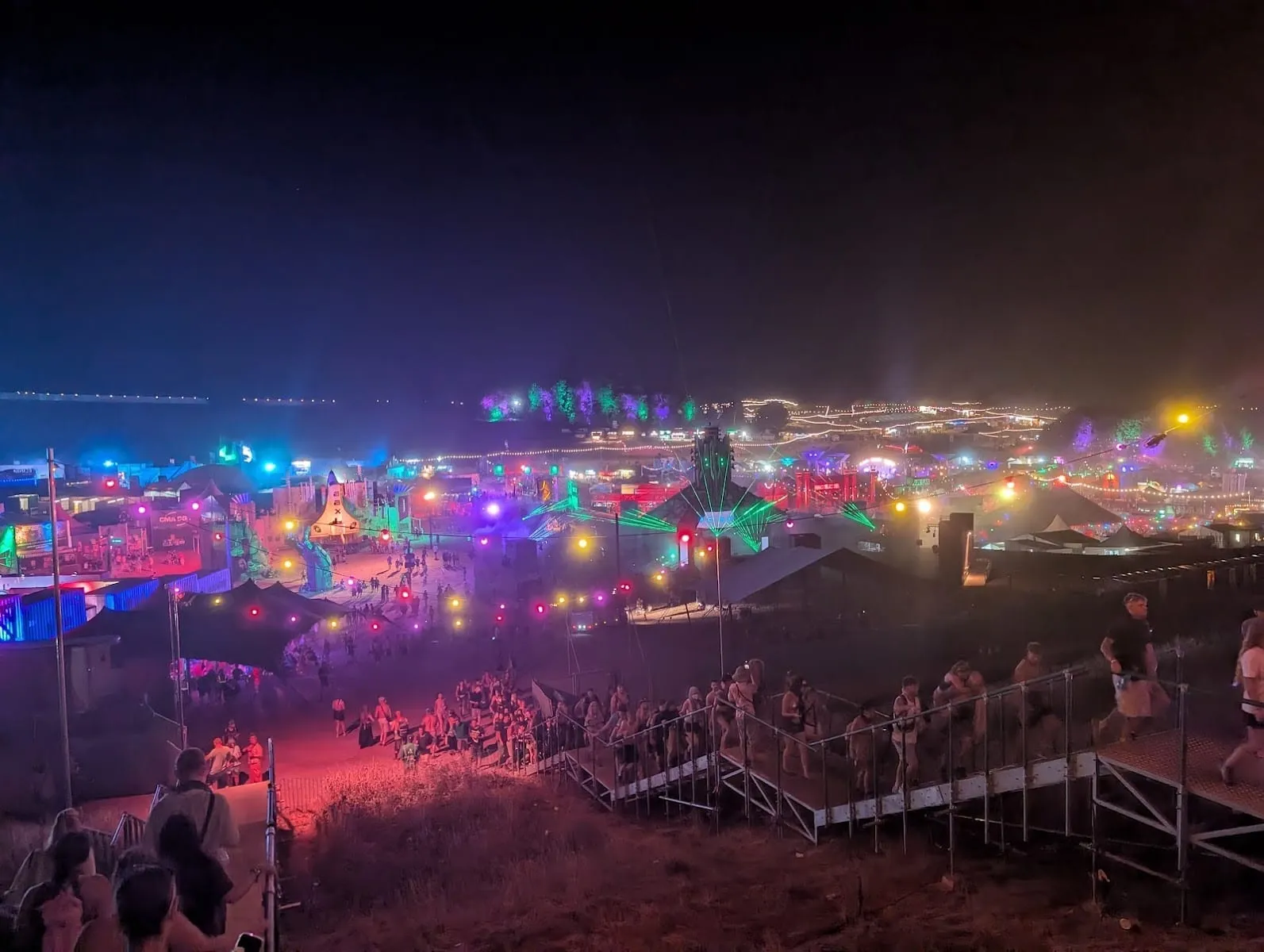 Boomtown's site at night seen from a hillside staircase, with festival-goers walking down towards lit-up stages, strings of coloured bulbs and green lasers