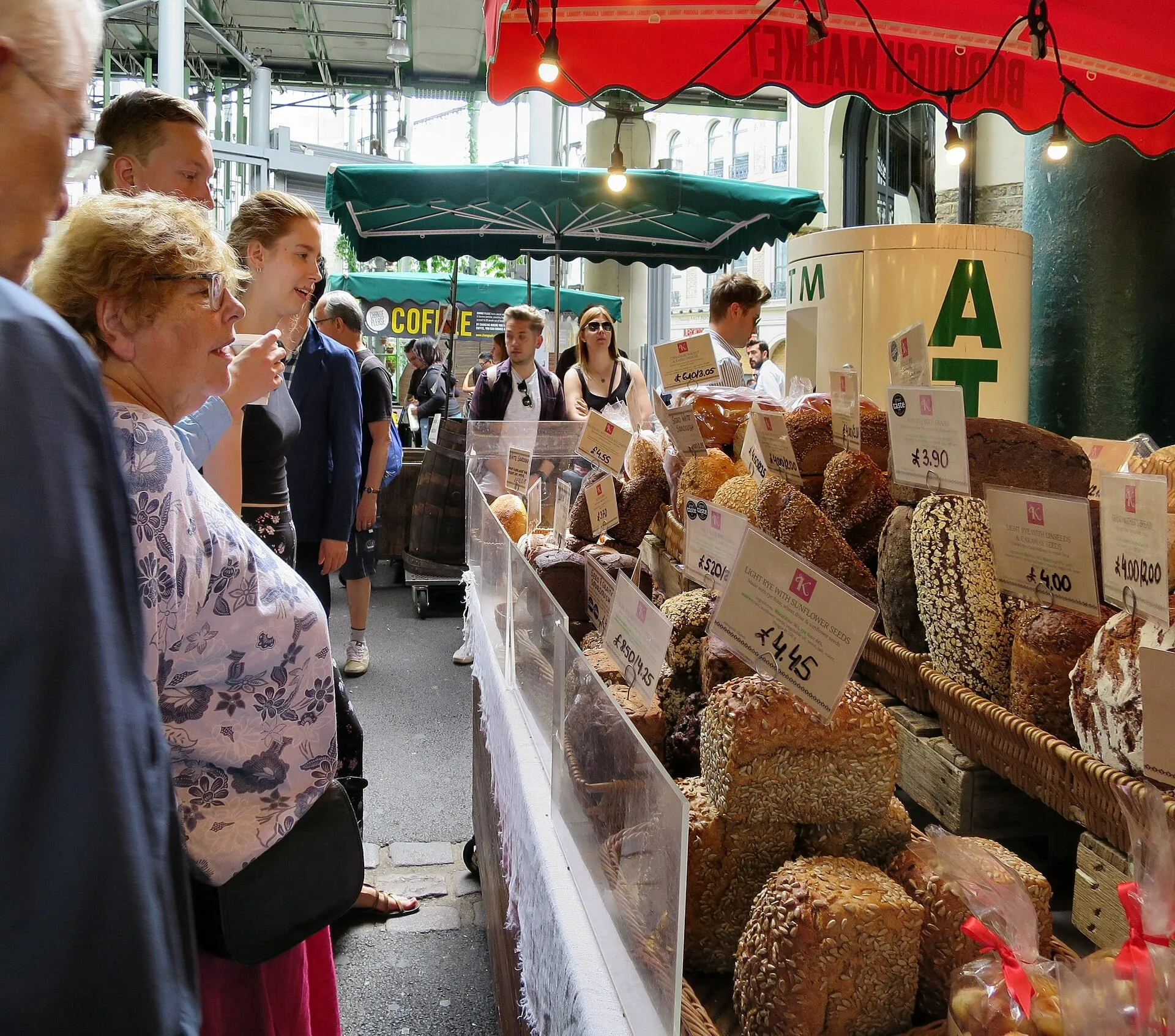 An artisan bakery stall at Borough Market displaying bread loaves with prices marked in pounds sterling and an ATM kiosk behind