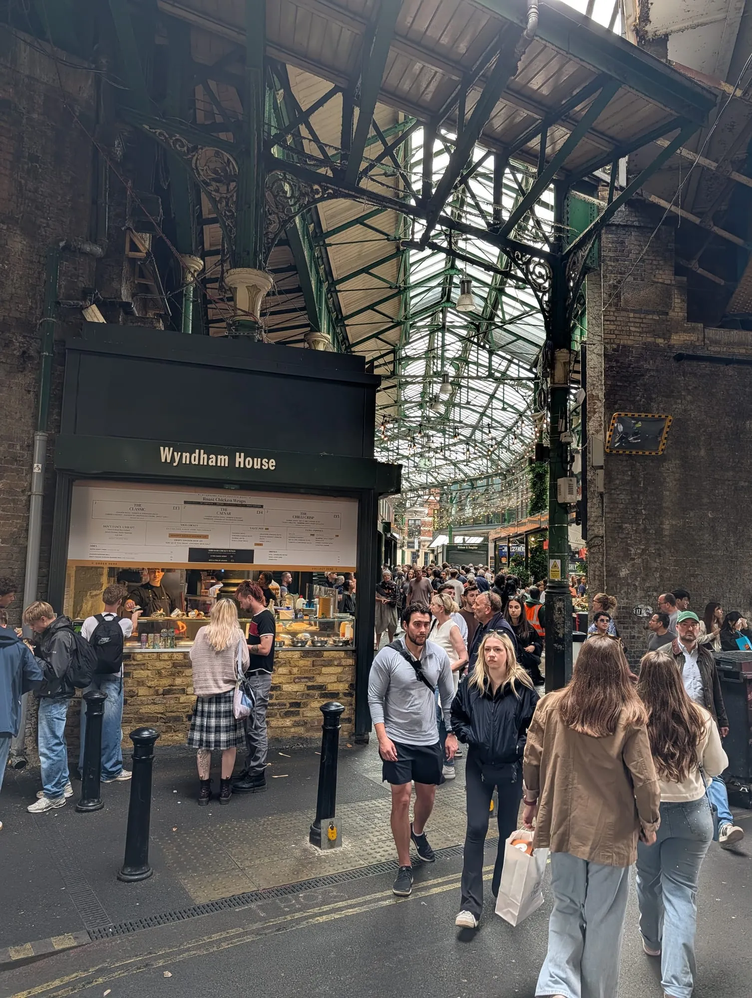 Crowds under the green ironwork and glass roof of Borough Market, a roast chicken wrap counter on the left with a queue at it