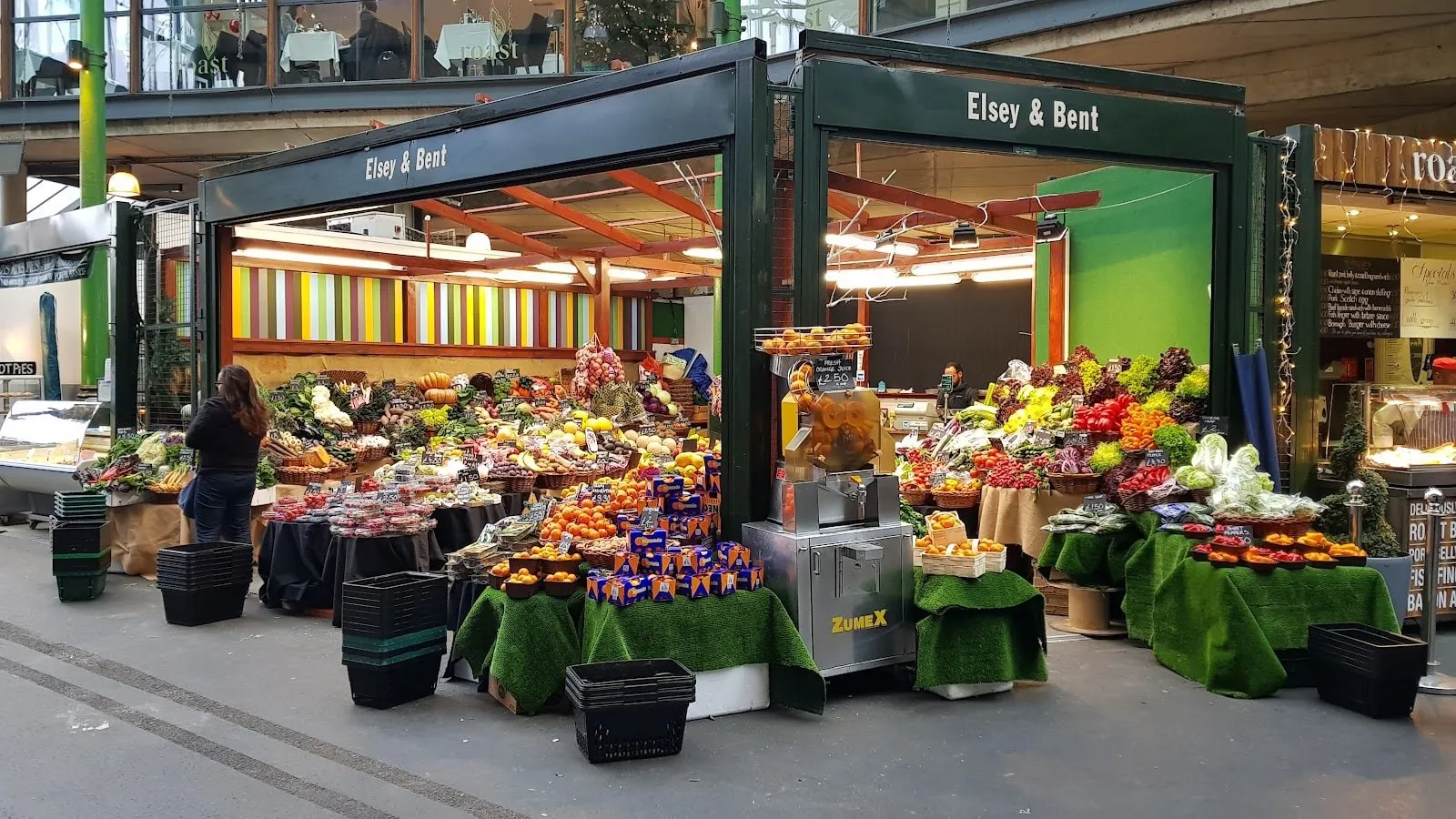 A greengrocer's stall signed Elsey & Bent in Borough Market, crates of fruit and vegetables on green cloths, a fresh orange juice press in the middle and a restaurant on the level above