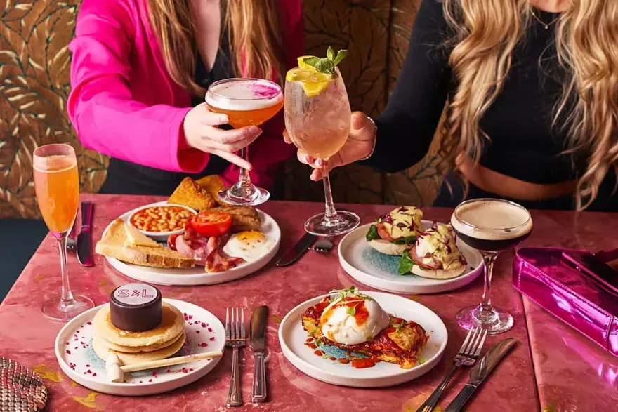 A bottomless brunch table: two people raising cocktails over a full English, eggs royale, pancakes and an espresso martini