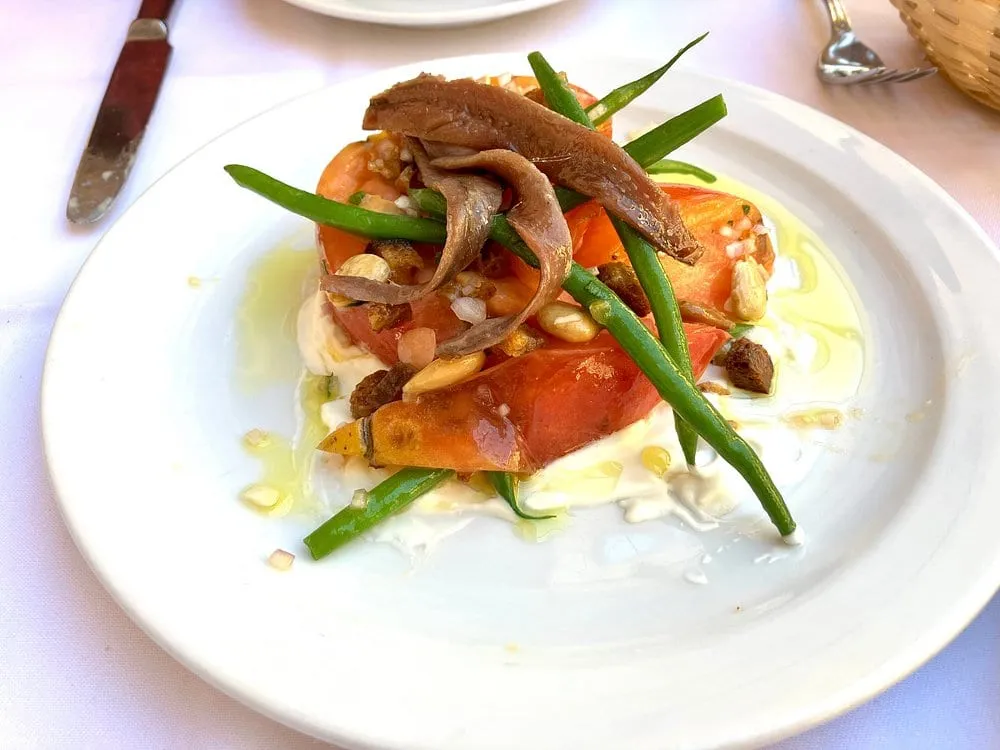 Tomatoes, green beans, anchovy fillets, almonds and croutons piled on a bed of cream on a white plate, on a white tablecloth
