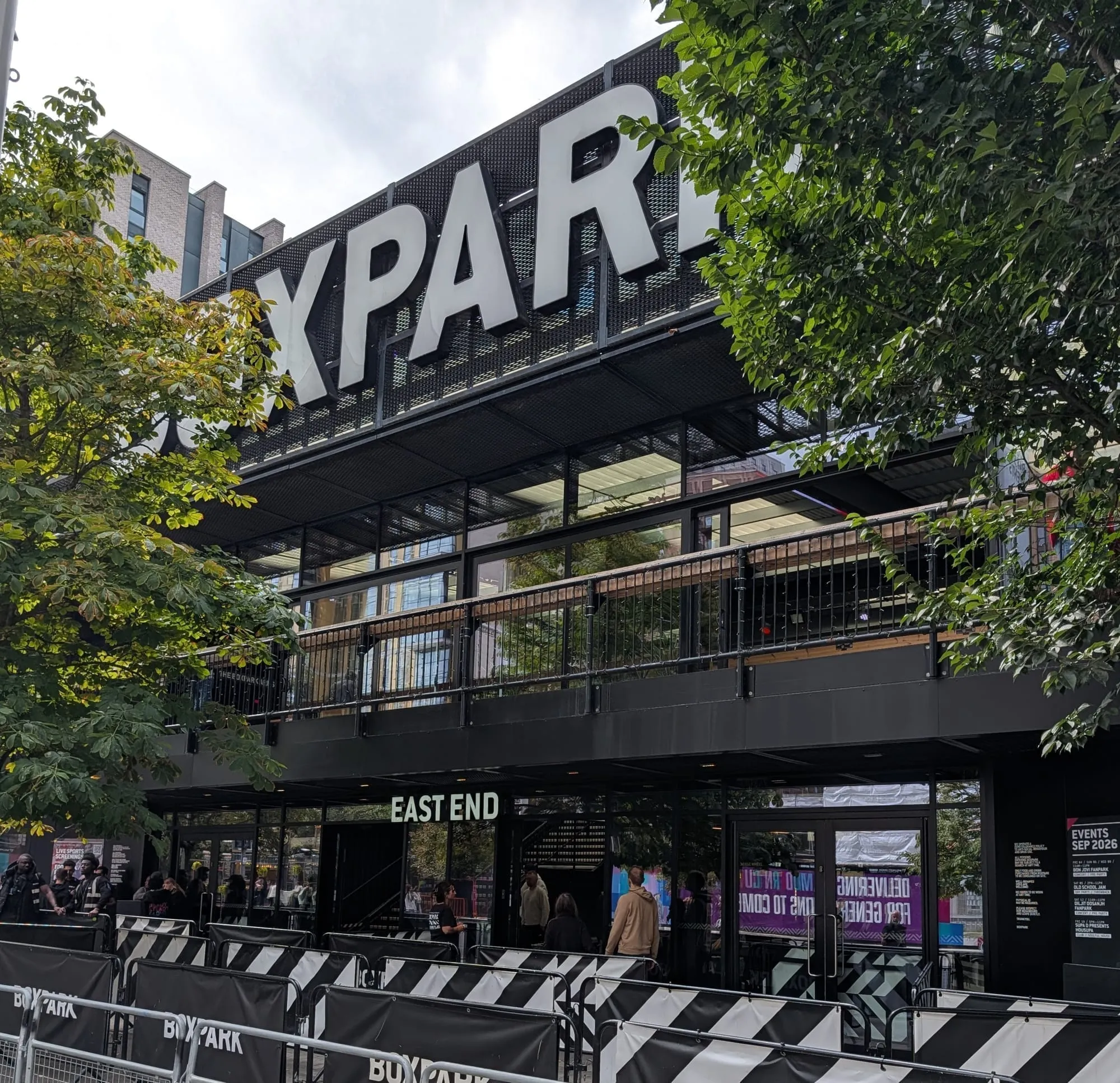 Boxpark Wembley's black frontage seen from below through trees, the white BOXPARK sign along the roofline, EAST END lettering over the doors and crowd barriers set out in front