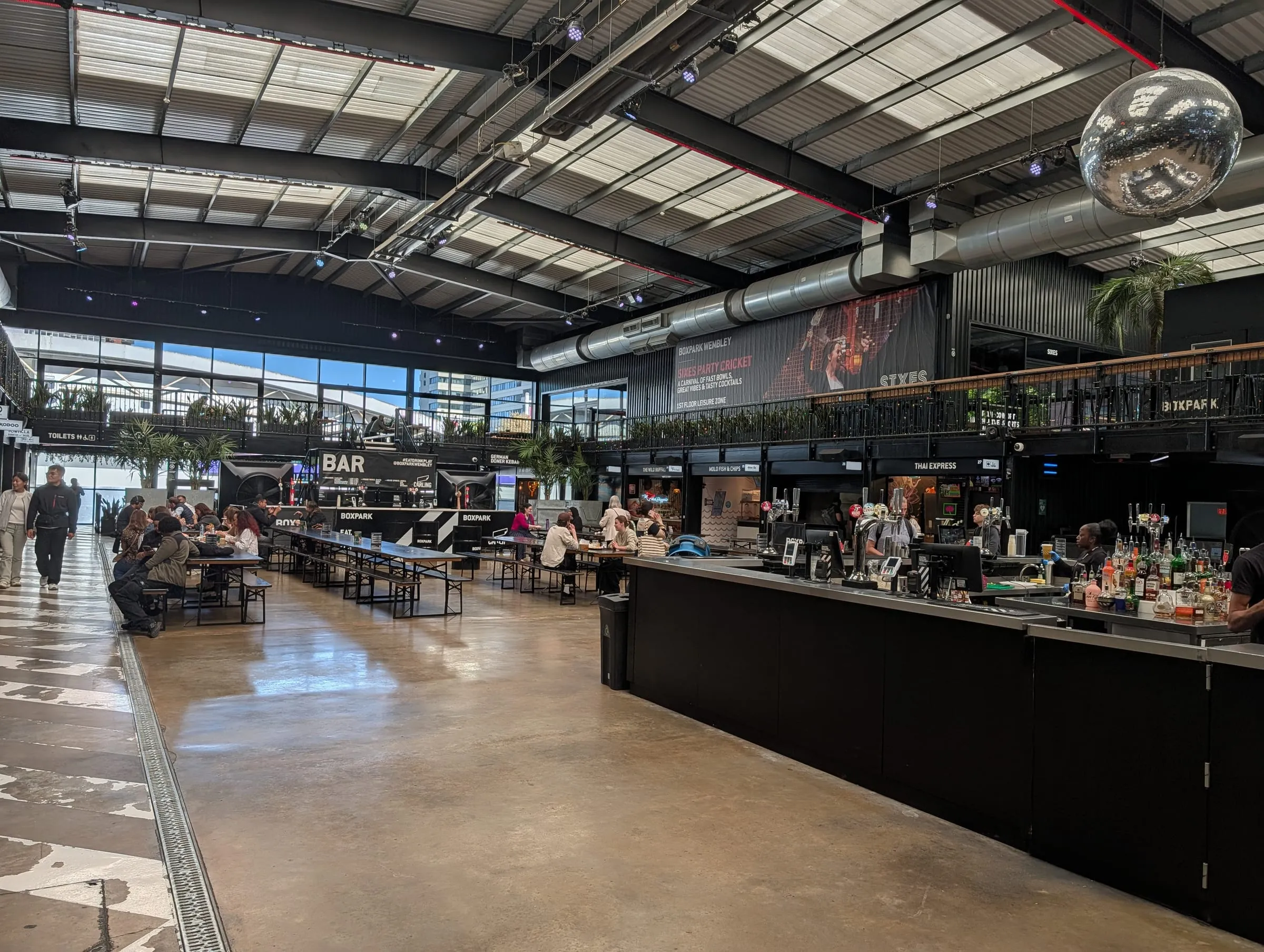 Inside Boxpark Wembley: a long black bar under a high warehouse roof with a mirror ball, and diners at long tables along the windows