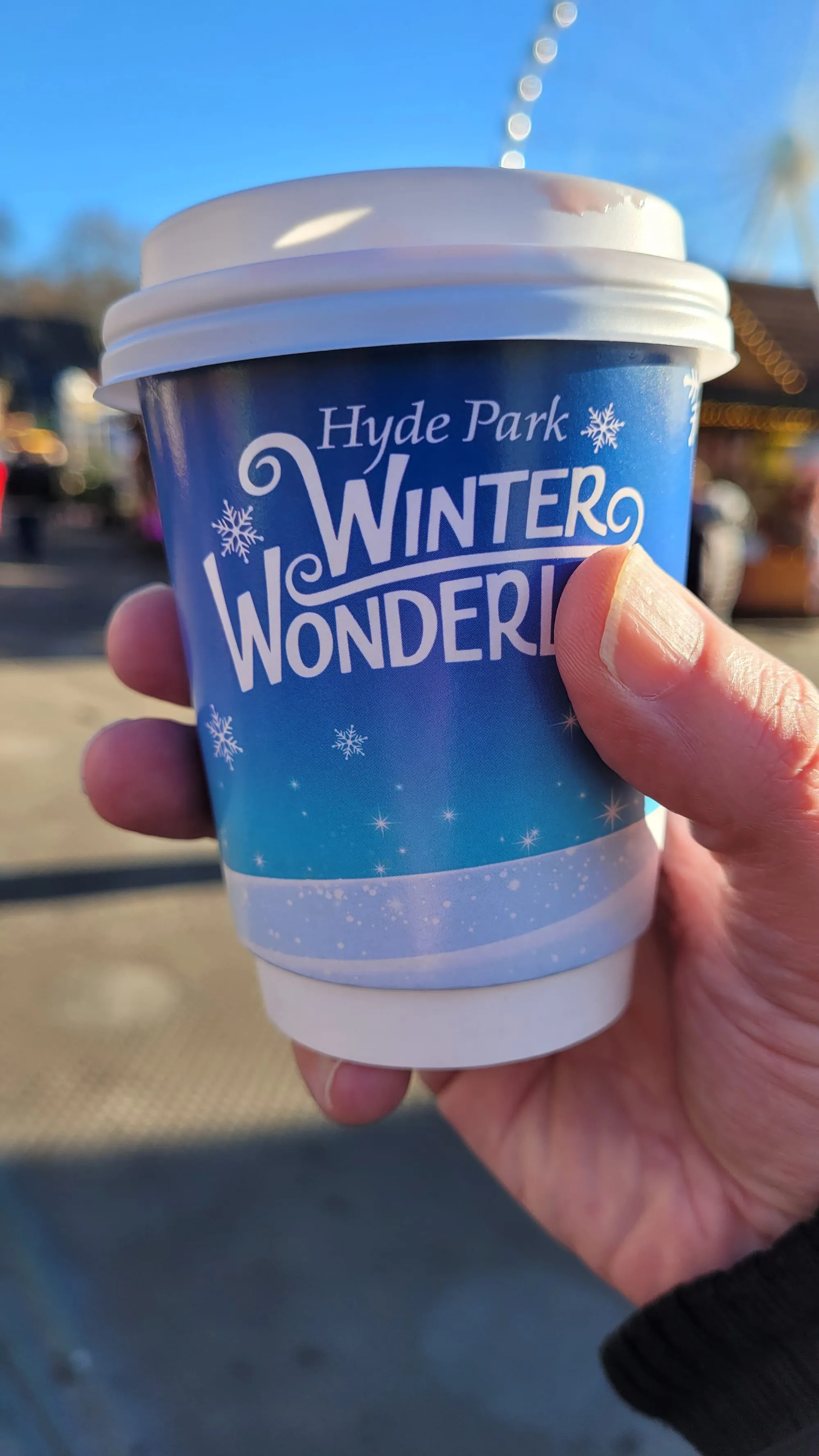 A hand holding a branded Hyde Park Winter Wonderland hot drinks cup, with the observation wheel out of focus behind